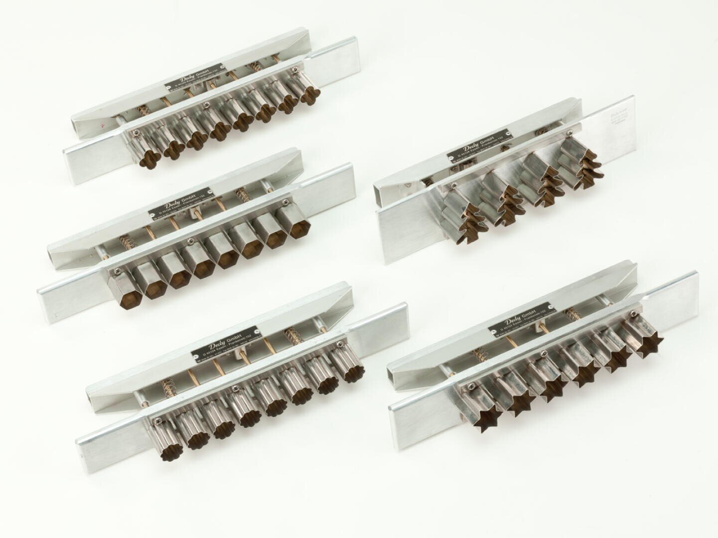 Fünf Metallextruderplatten mit verschieden geformten Löchern, darunter Sterne, Herzen, Sechsecke und Bäume, sind auf einer weißen Fläche angeordnet. Die Platten werden für die Formung von Teig oder ähnlichen Materialien verwendet.