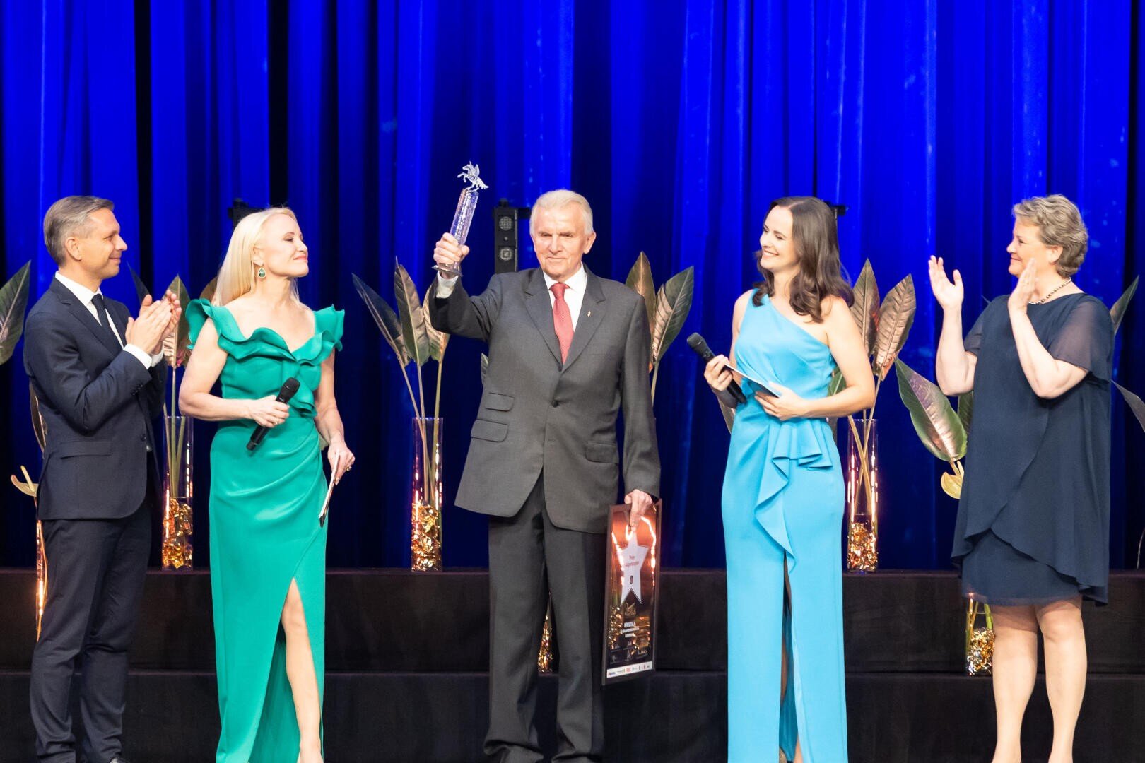 Ein älterer Mann im Anzug hält auf der Bühne eine Trophäe hoch, umgeben von vier formell gekleideten Personen. Blaue Vorhänge und Pflanzen schmücken den Hintergrund, während die anderen lächeln, klatschen und zu ihm blicken.