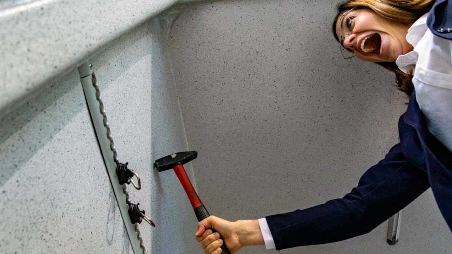 Eine Frau in Geschäftskleidung schwingt enthusiastisch einen Hammer auf einen Schlüsselbund, der in einer Wand steckt, den Mund vor Aufregung oder Entschlossenheit geöffnet.