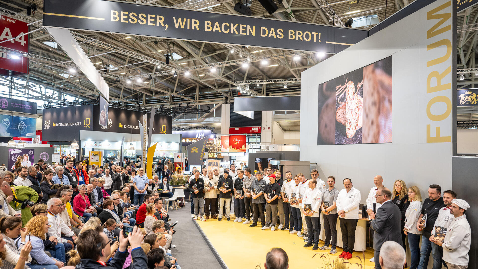 Ein großes Publikum beobachtet eine Gruppe von Bäckern, die bei einer Backveranstaltung in einer Kongresshalle auf der Bühne stehen. Auf einem Banner über der Bühne steht "Besser, wir backen das Brot!", und auf dem Bildschirm ist ein großes Brotbild zu sehen.