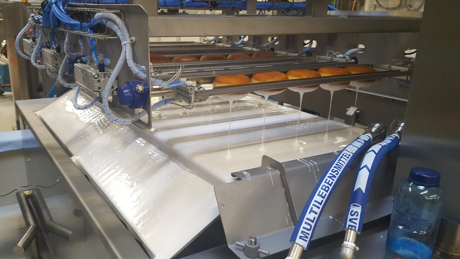 Reihen von Donuts auf einem Förderband laufen unter einer Maschine hindurch, die sie mit weißer Glasur übergießt, wie in einer industriellen Lebensmittelverarbeitung. Um den Glasurbereich herum sind blaue Schläuche und Metallmaschinen zu sehen.