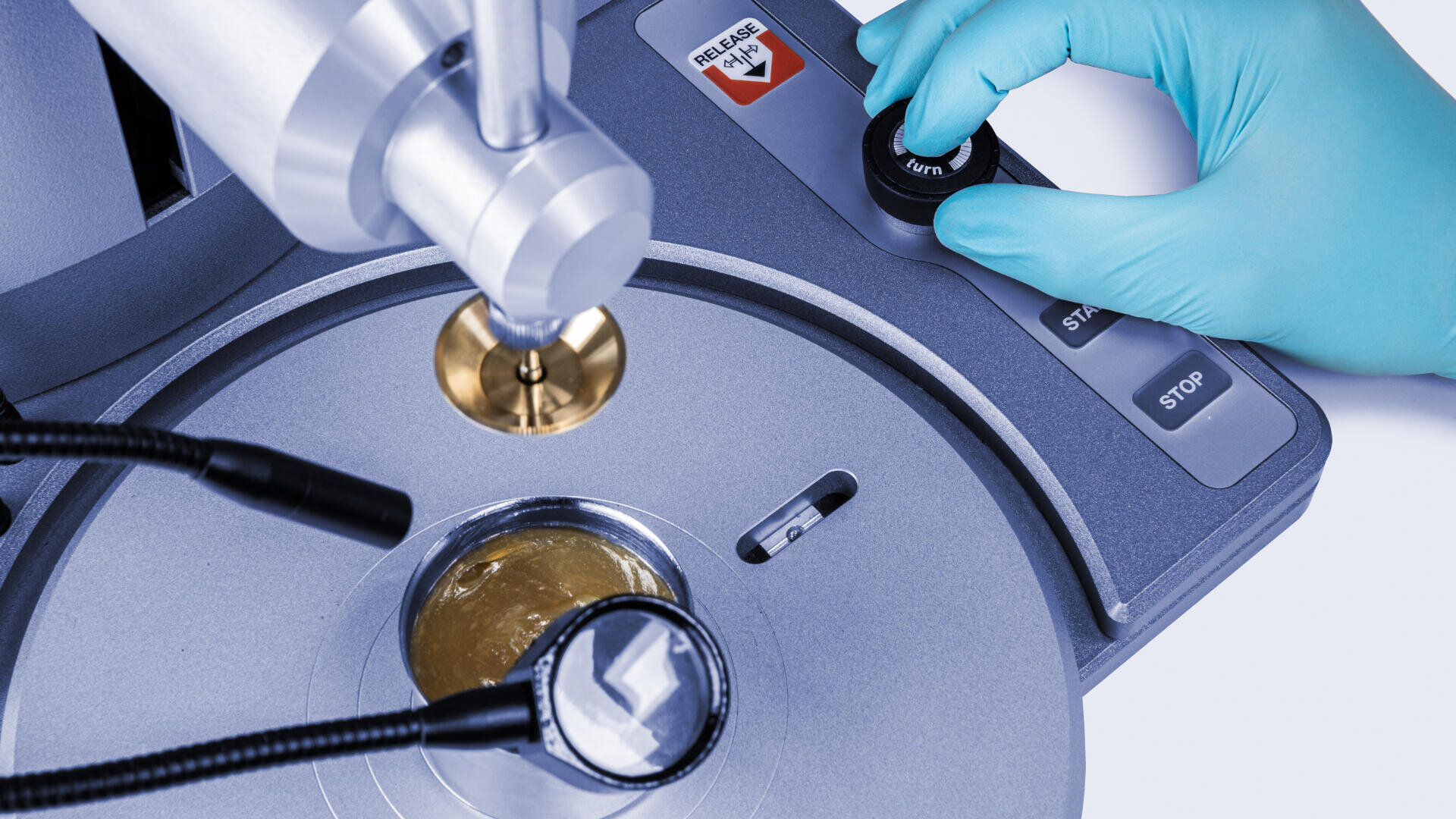 A gloved hand operates a control panel of a laboratory instrument with metallic and glass components used for scientific analysis or material testing.