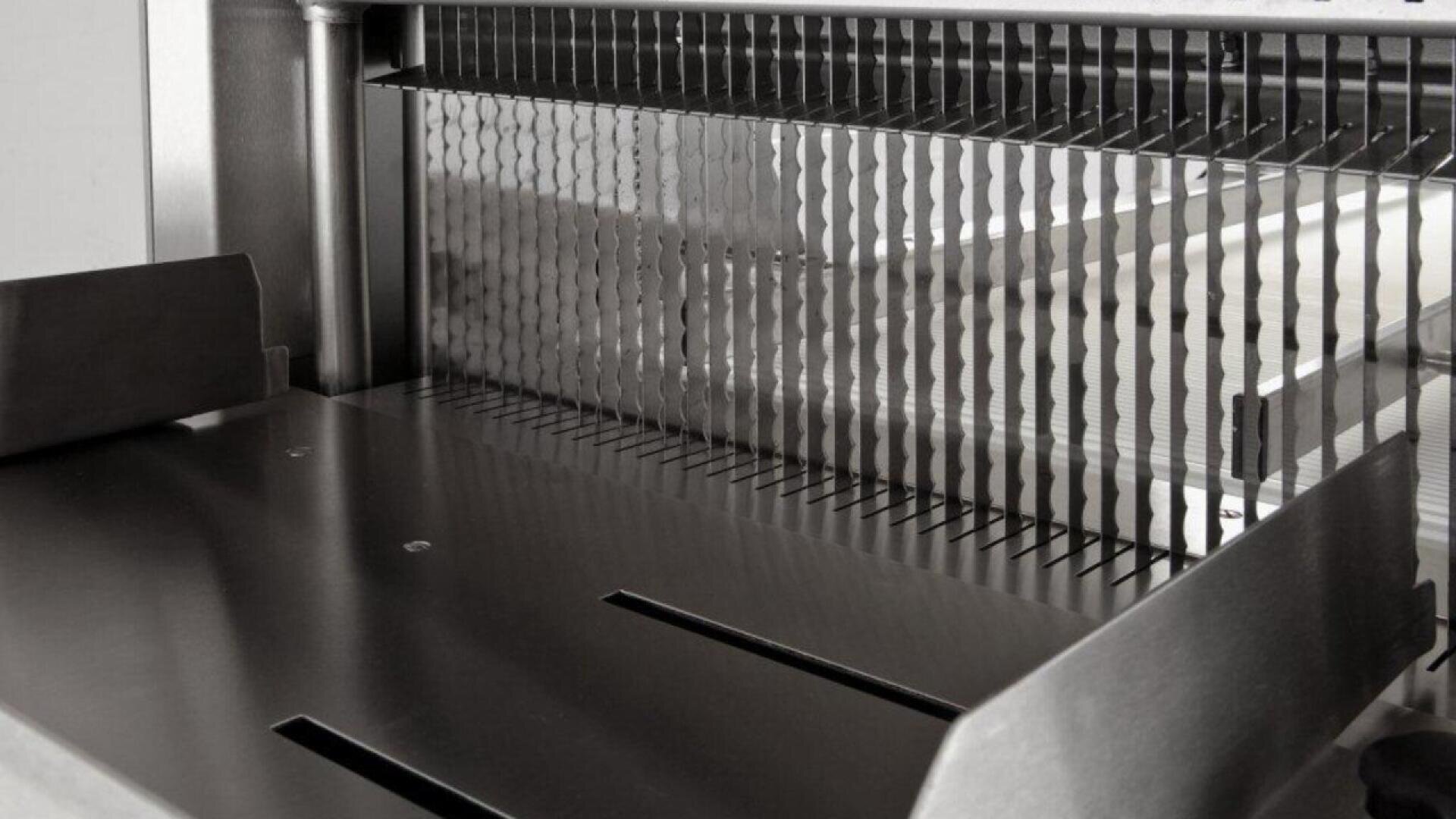 Close-up view of a commercial bread slicer with multiple evenly spaced, wavy metal blades designed for cutting loaves into uniform slices. The surrounding area appears to be made of stainless steel.