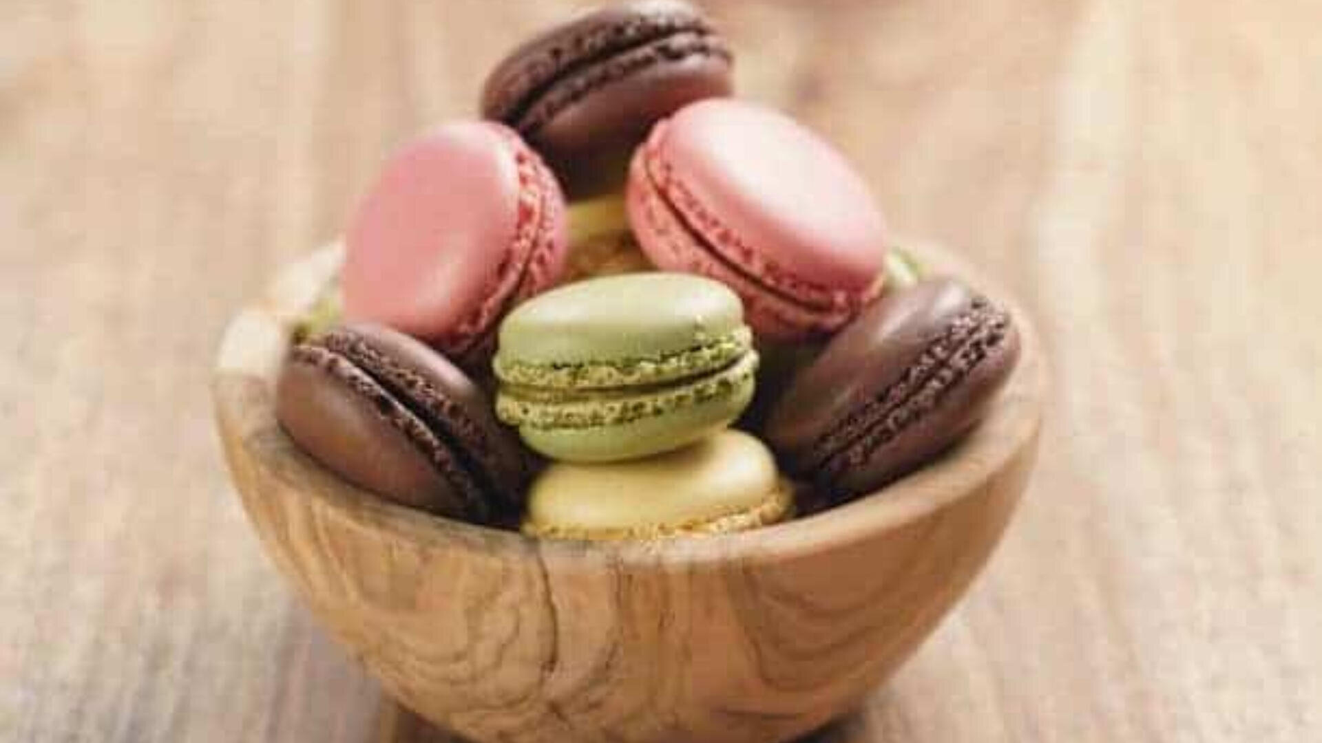 Auf einer hölzernen Unterlage steht eine Holzschale, gefüllt mit verschiedenen bunten Macarons, darunter rosa, grün, gelb und braun.