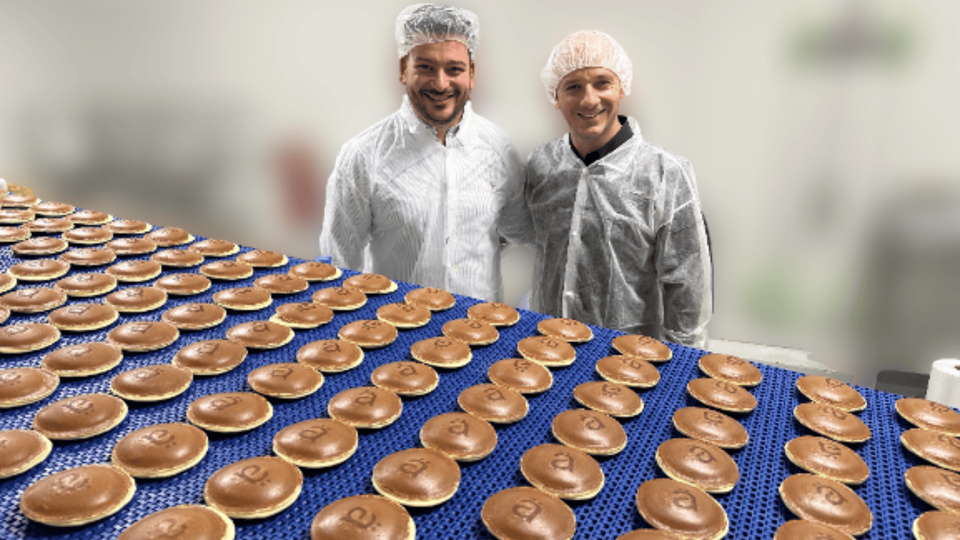 Two people in white hairnets and lab coats stand in a factory next to a conveyor belt covered with rows of round, brown baked goods. The background is blurred, focusing on the people and the pastries.
