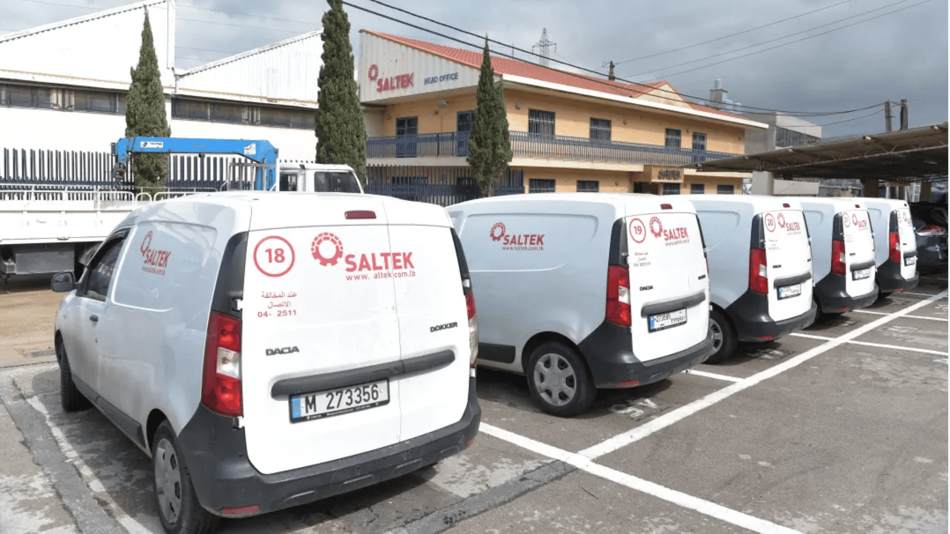 Five white vans with SALTEK branding are parked in a row in a lot outside a building with the same logo on the facade. The scene includes trees and an overcast sky.