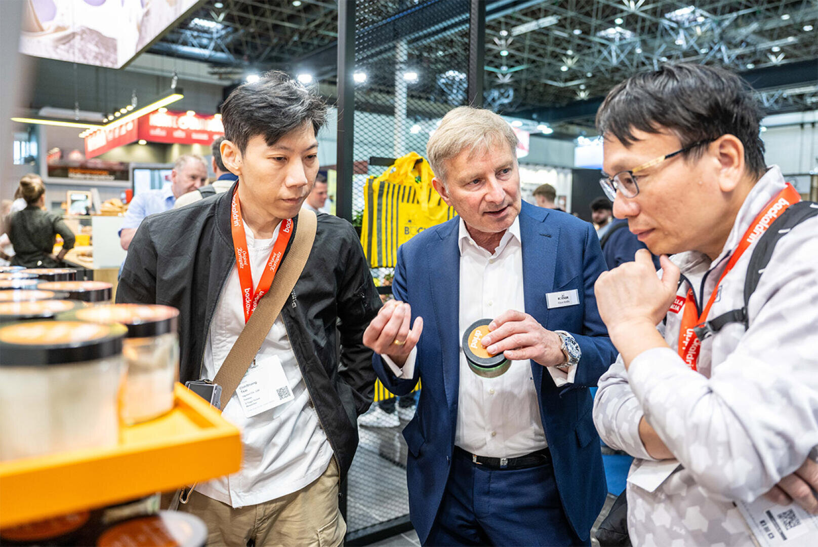 Ein Mann im blauen Anzug erklärt zwei Männern mit roten Schlüsselbändern an einem Messestand ein Produkt. Im Hintergrund sind Regale und andere Besucher zu sehen.