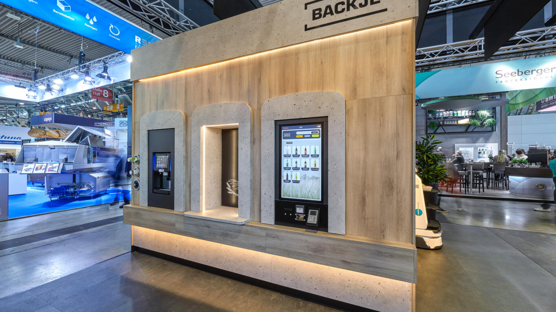 Ein moderner Selbstbedienungs-Verkaufskiosk mit digitalen Touchscreens und Ausgabefenstern in einer Messeumgebung mit Messeständen und Geräten im Hintergrund.