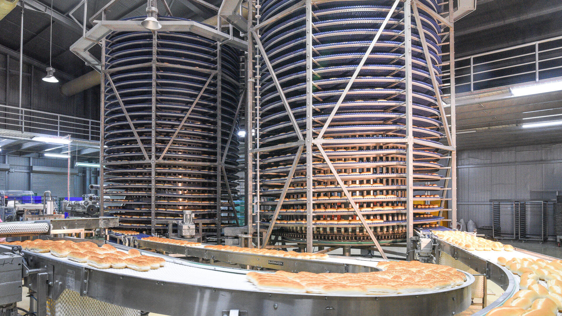 Loaves of bread move along a conveyor belt in a large industrial bakery, with tall spiral cooling racks stacked with bread in the background. The setting is clean, bright, and modern.