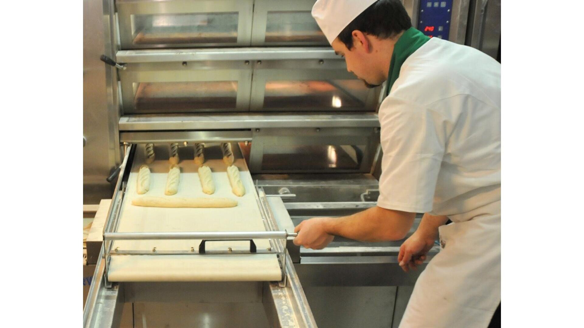A baker in a white uniform and hat is placing uncooked baguettes onto a baking tray using a sliding wooden peel in front of an industrial oven.