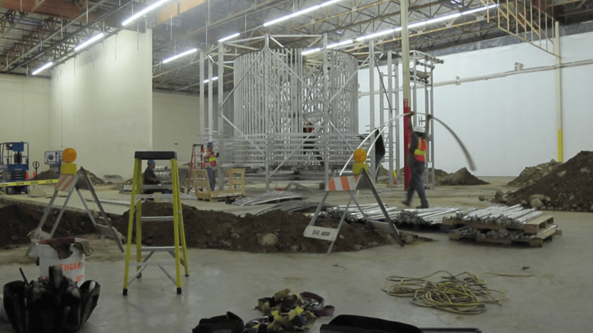 Construction workers in safety vests are building metal framework structures inside a large, unfinished indoor space. There are scattered tools, cables, dirt piles, and safety barriers around the work area.
