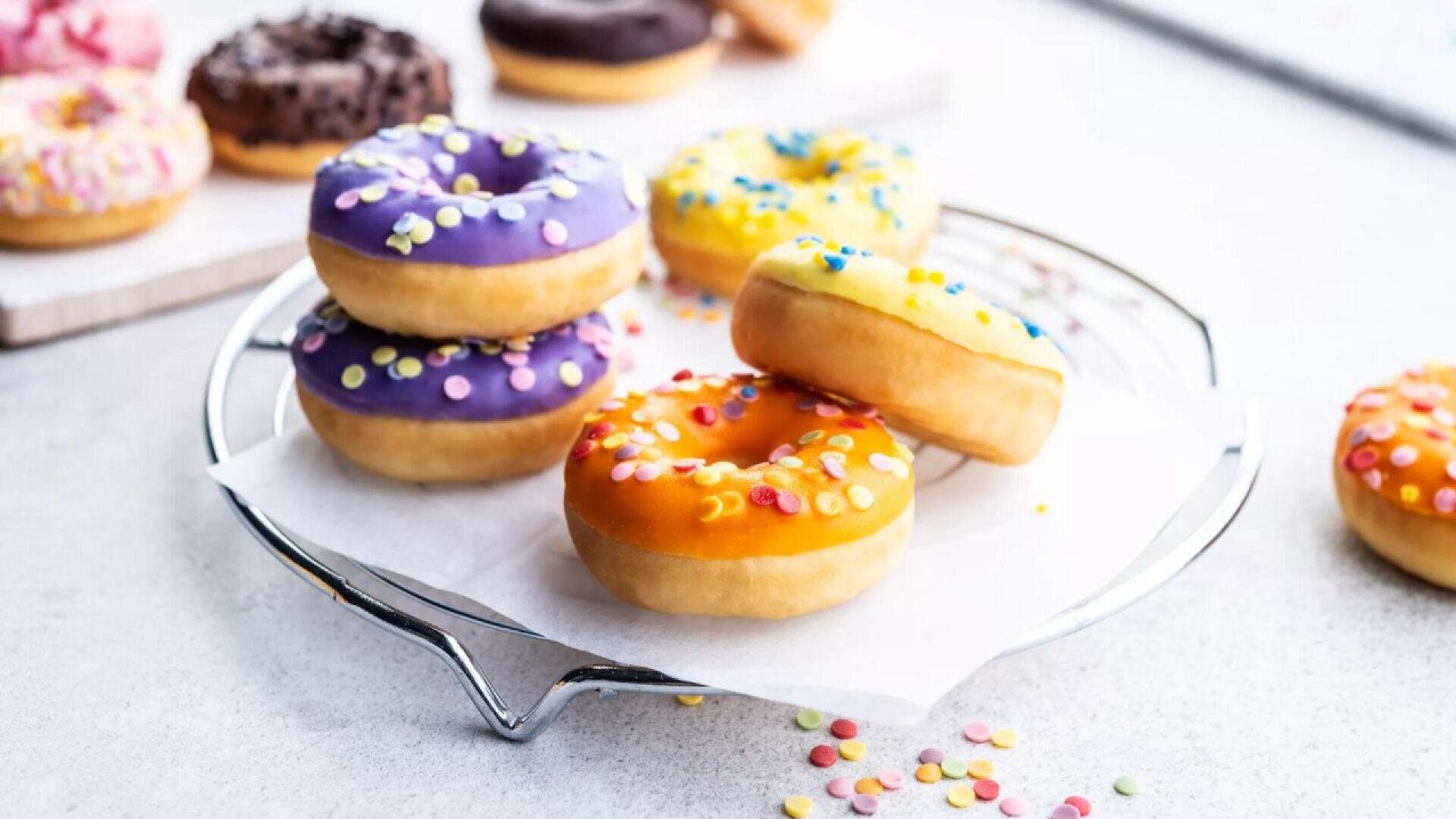 A plate of colorful, frosted donuts with sprinkles sits on parchment paper. Donuts are topped with purple, yellow, and orange icing. More assorted donuts are blurred in the background.