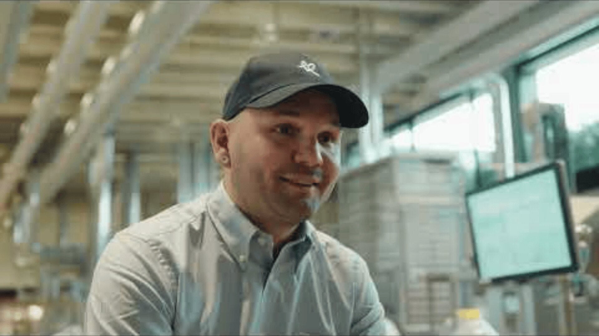 Ein lächelnder Mann mit Baseballmütze und zugeknöpftem Hemd sitzt in einem Raum, in dem im Hintergrund Industrieanlagen und ein Computermonitor zu sehen sind. Die Umgebung scheint ein moderner Arbeitsplatz oder eine Fabrik zu sein.