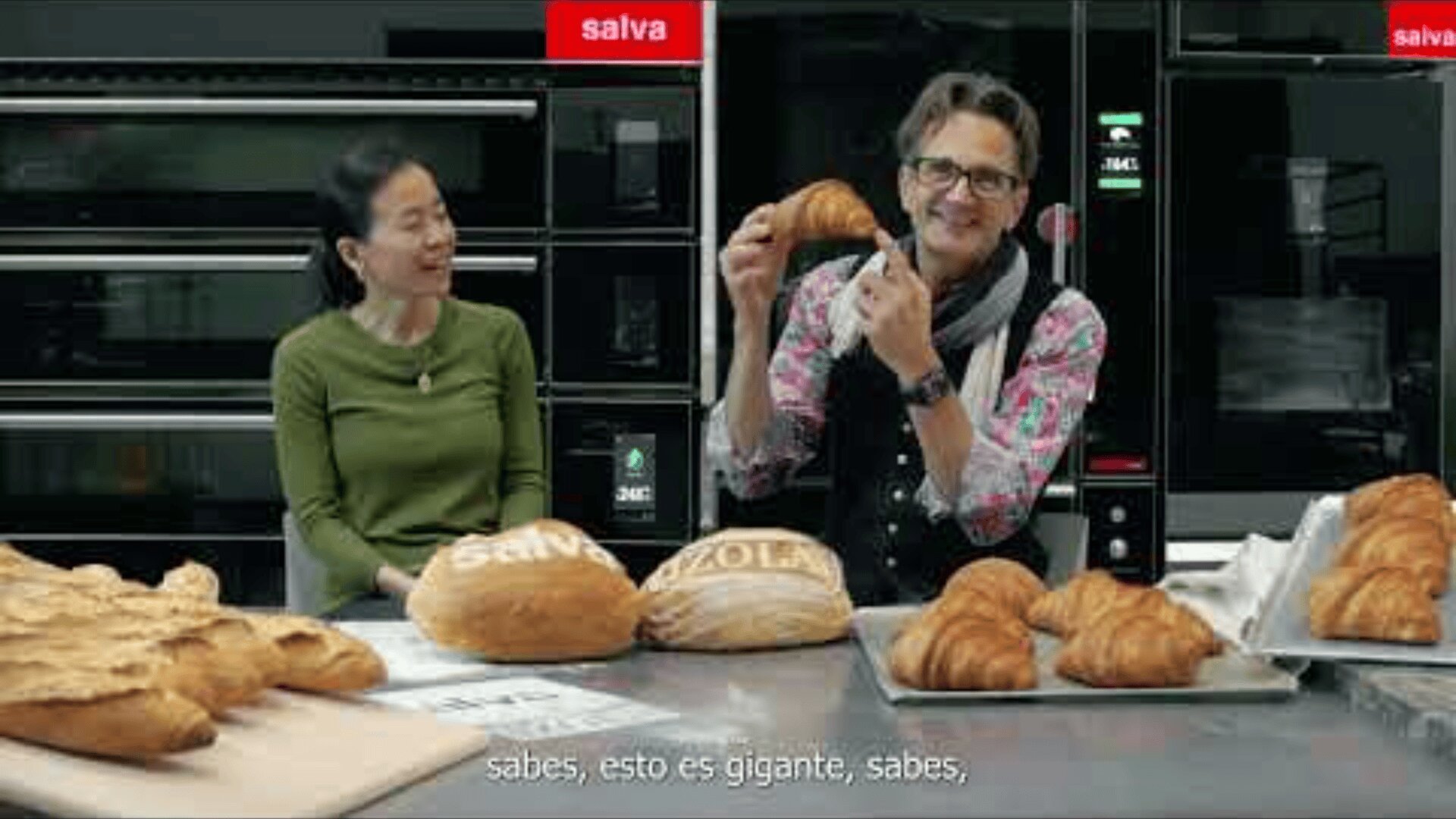 Two people sit at a table with various breads and pastries in front of them. The man holds up a large croissant and smiles, while the woman looks at him and smiles. Industrial ovens are visible in the background.