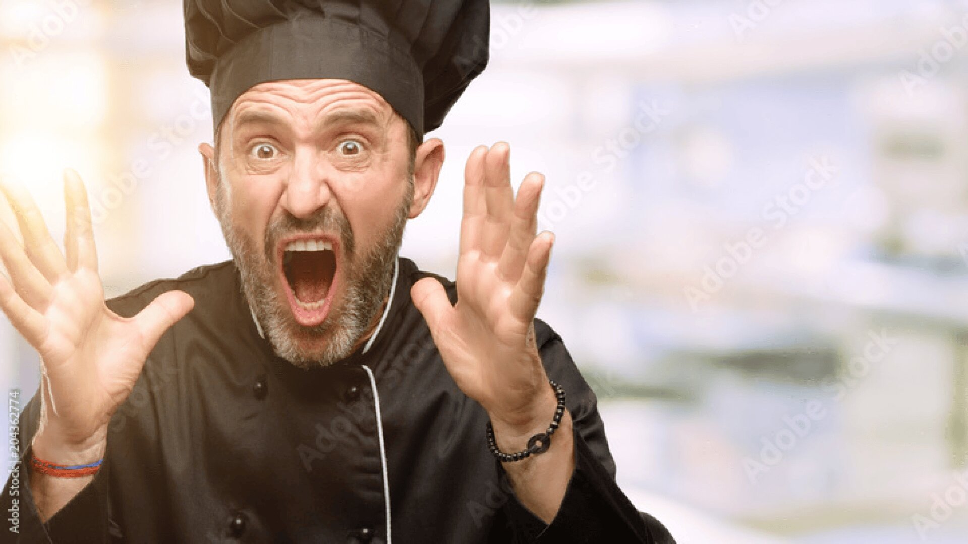 Ein männlicher Koch in schwarzer Uniform und mit Mütze steht mit schockiertem Gesichtsausdruck, weit aufgerissenem Mund, großen Augen und erhobenen Händen in einem Raum. Der Hintergrund ist unscharf.