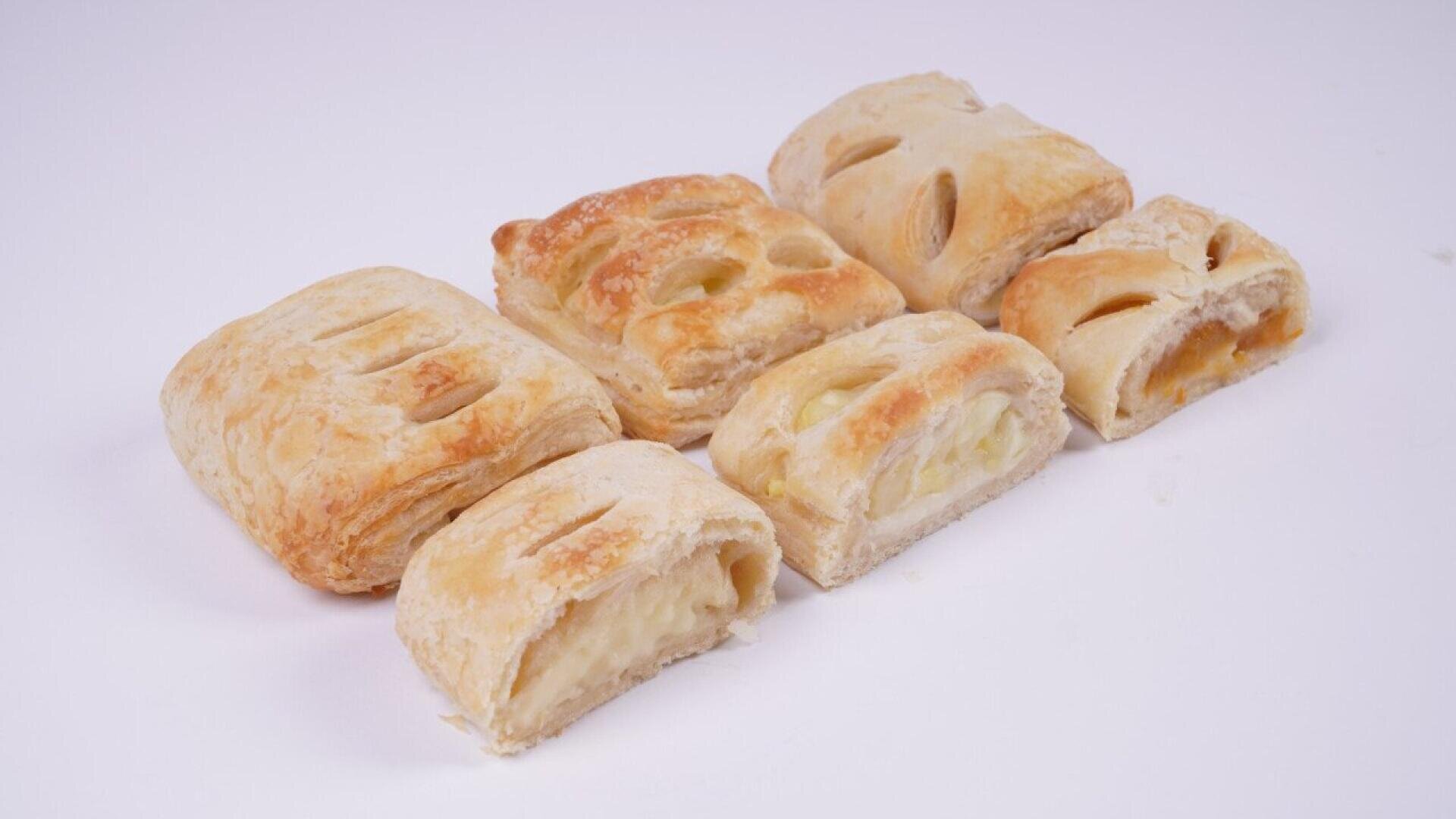 Six pieces of golden-brown, rectangular puff pastries with visible flaky layers are arranged on a plain white background. Some pastries have small slits on top, revealing a light cream or fruit filling inside.