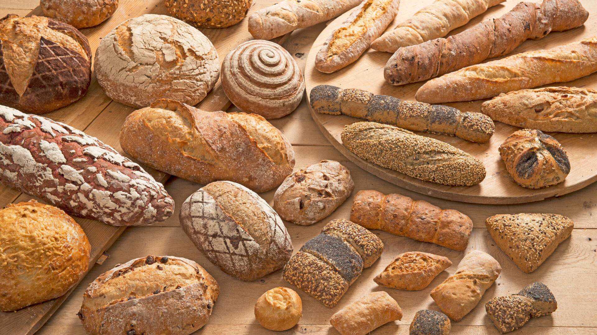 Assorted loaves and rolls of artisan bread in various shapes and sizes are arranged on wooden boards and a tabletop, showcasing different crust textures, patterns, and seeds.