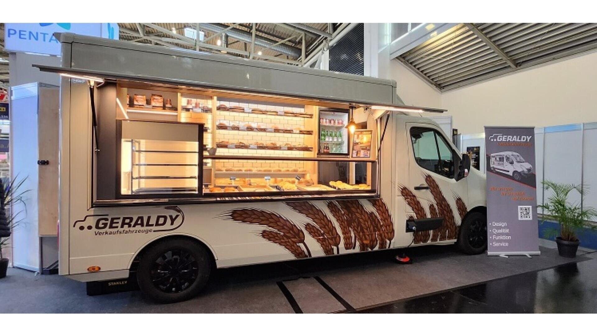 A modern food truck with an open service window displays shelves filled with bread and pastries. The truck has wheat graphics on the side and is parked indoors next to a sign and some potted plants.