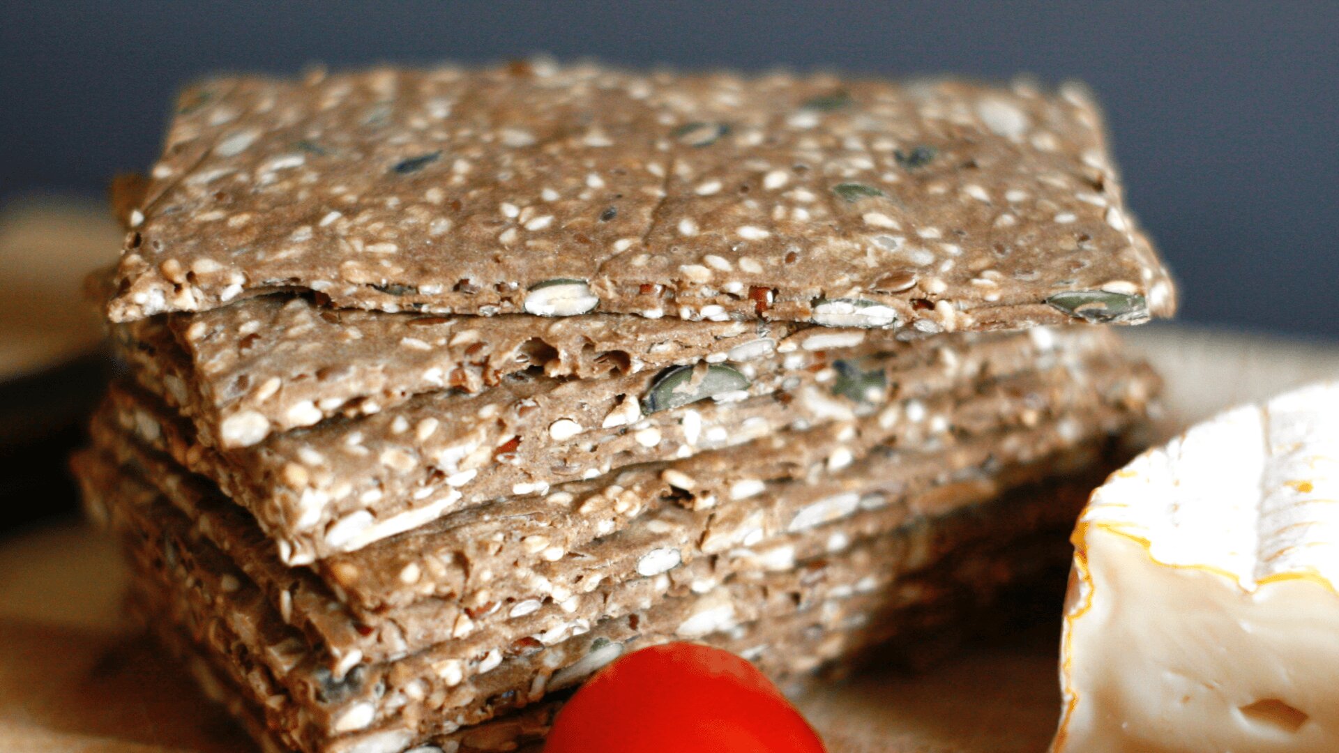 A stack of rectangular, seeded crispbread sits on a wooden surface, next to a wedge of soft cheese and a cherry tomato.