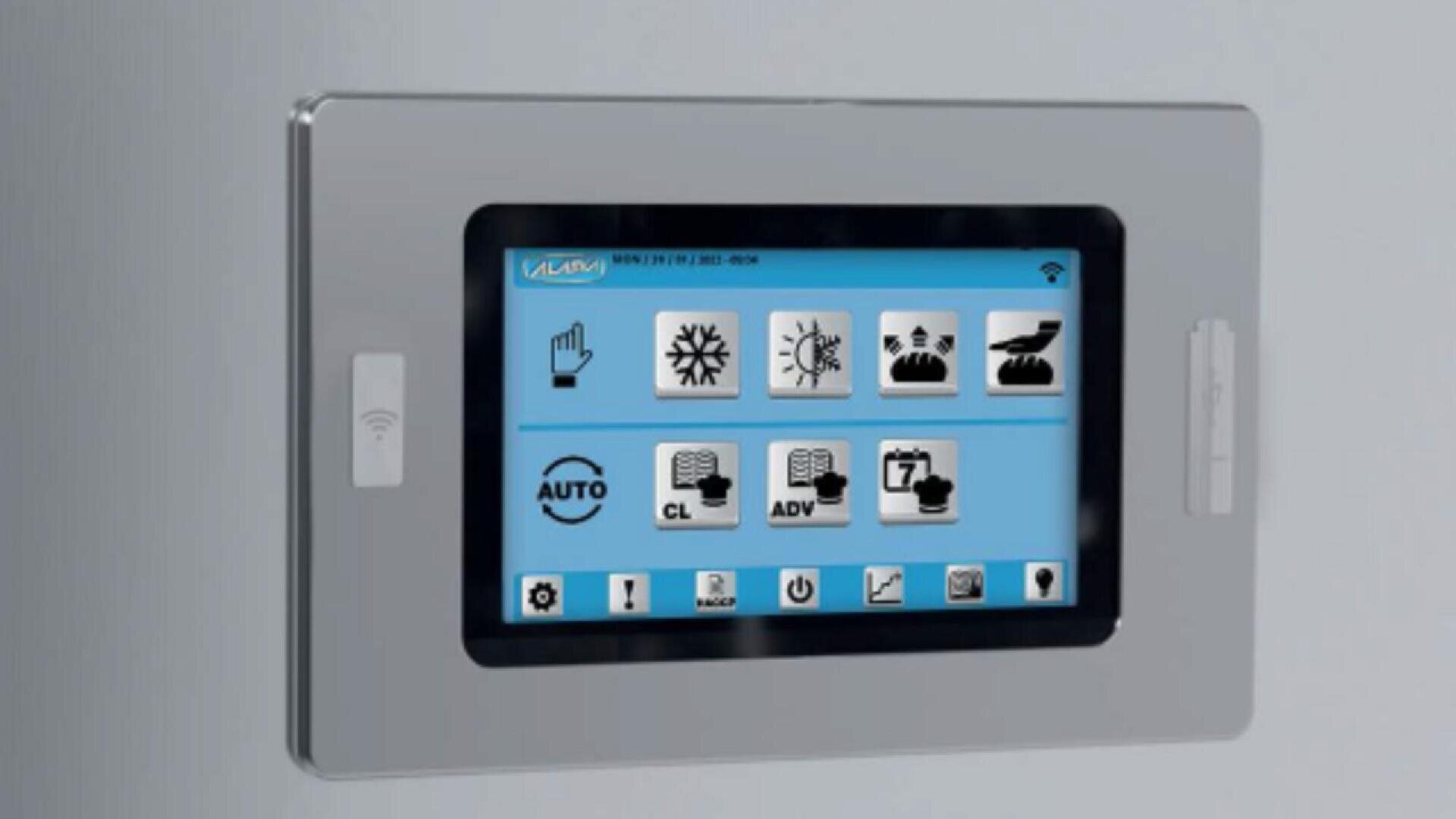 A wall-mounted digital control panel with a touchscreen display showing various icons for home automation functions, such as lighting, temperature, and scheduling, on a blue background.