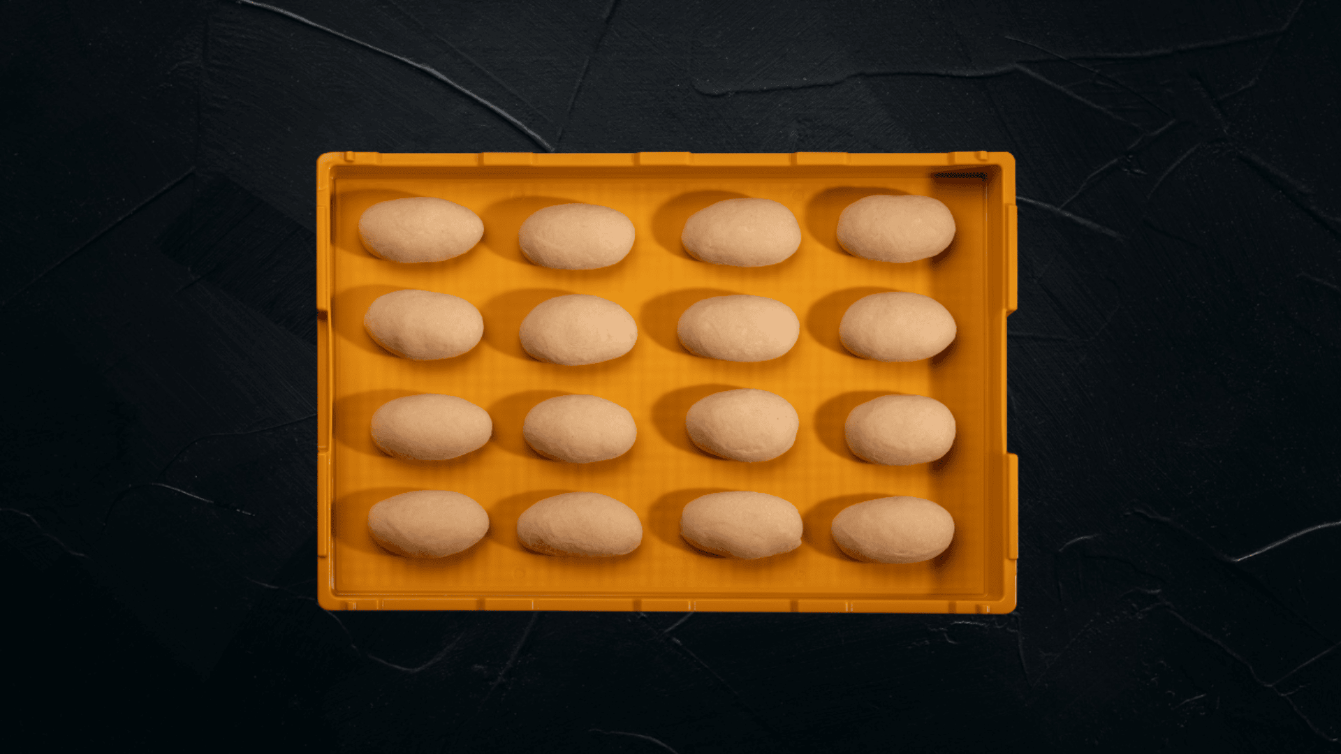 Auf einem gelben Tablett befinden sich fünfzehn gleichmäßig verteilte, ovale Teigkugeln auf einer dunklen, strukturierten Oberfläche.