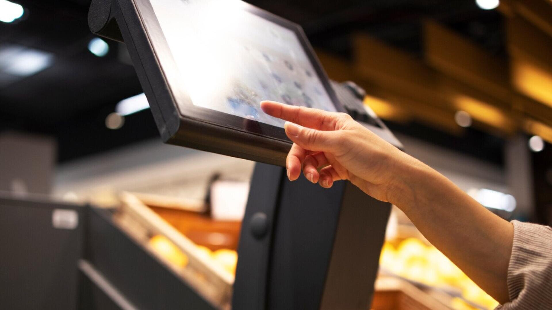 A person’s hand touches the screen of a self-service kiosk in a store, with blurred shelves and products visible in the background.
