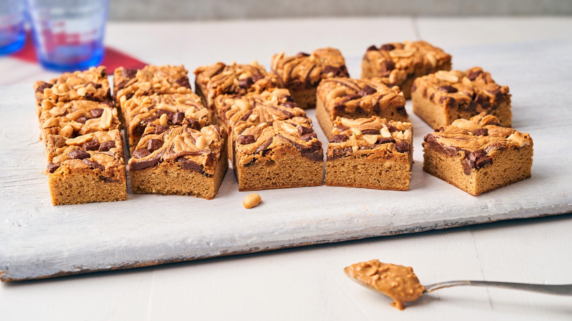 Rectangular peanut butter blondies topped with chopped peanuts and chocolate, arranged in rows on a light wooden board. A spoon with a dollop of peanut butter is placed in the foreground.