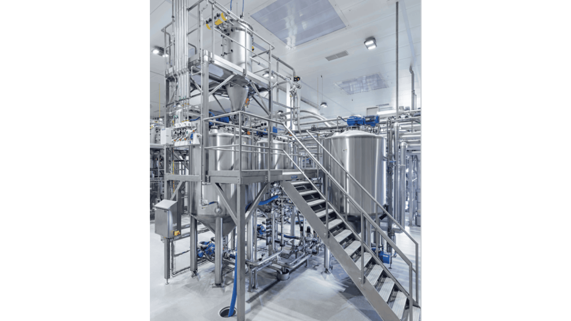 A modern industrial facility interior showing large stainless steel tanks, metal walkways, ladders, and pipes, with bright overhead lighting and a clean, organized environment.