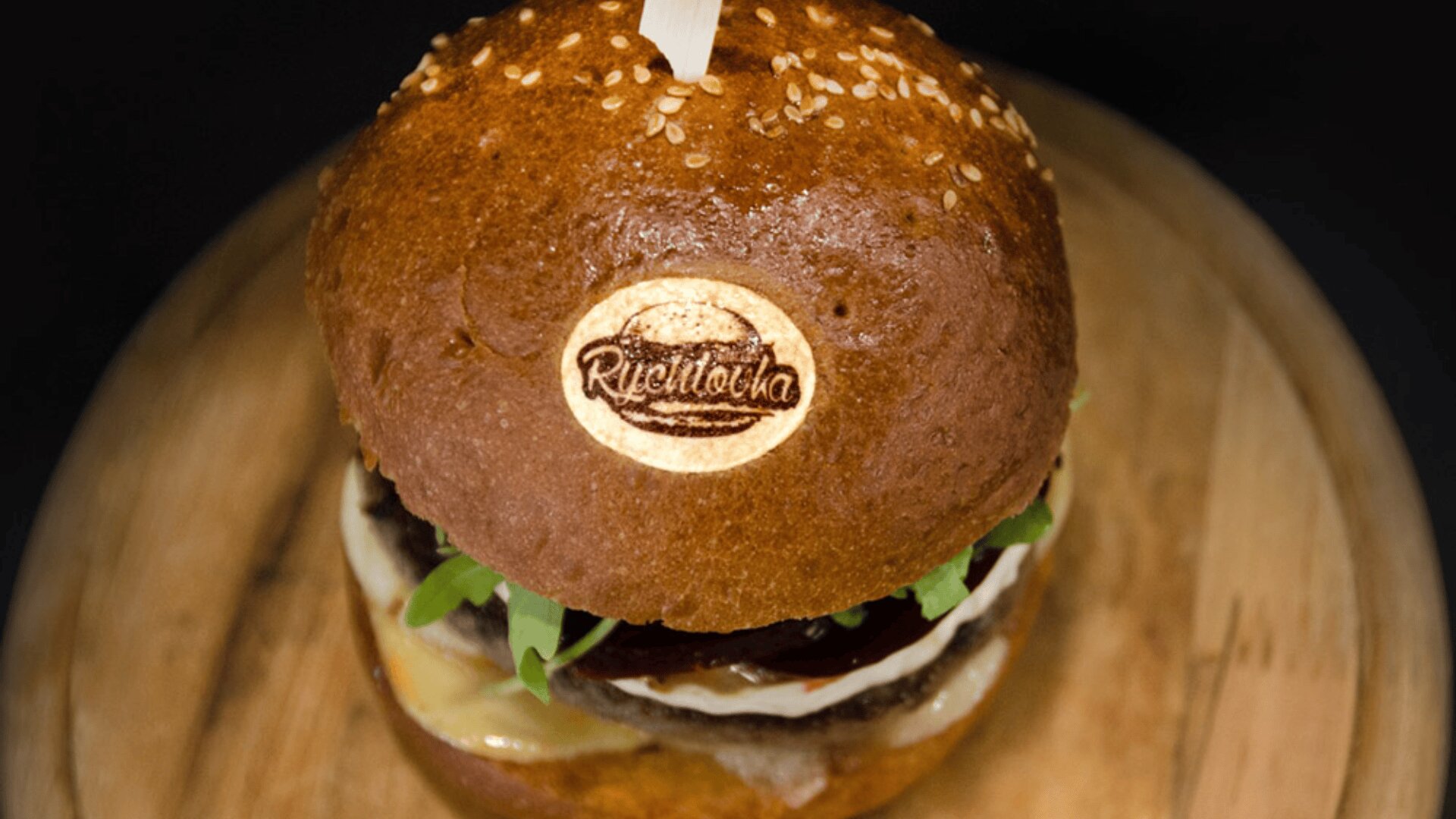 A close-up of a gourmet burger on a wooden board, featuring a toasted sesame bun branded with a Rijekačka logo, fresh greens, melted cheese, and a wooden skewer holding it together.