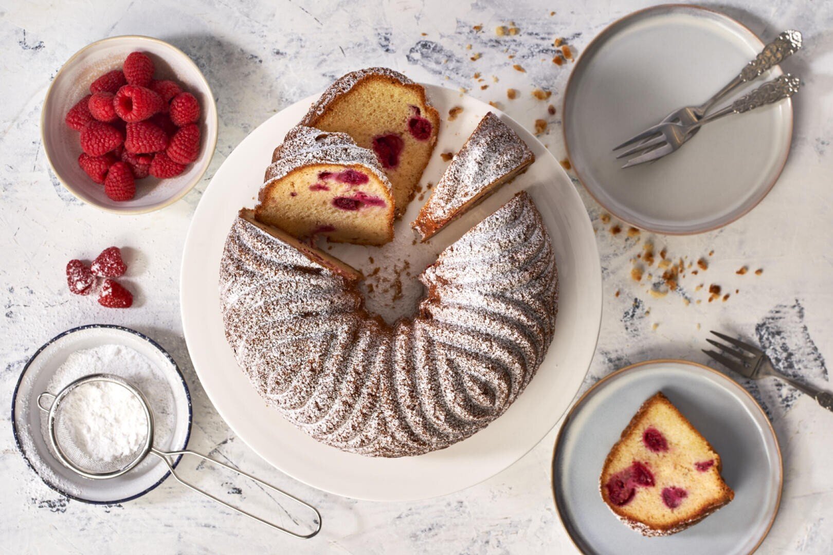 A Bundt cake with raspberries inside is sliced on a white plate. Two slices are served on gray plates with forks. Fresh raspberries and powdered sugar in bowls are nearby. Crumbs are scattered on the white surface.