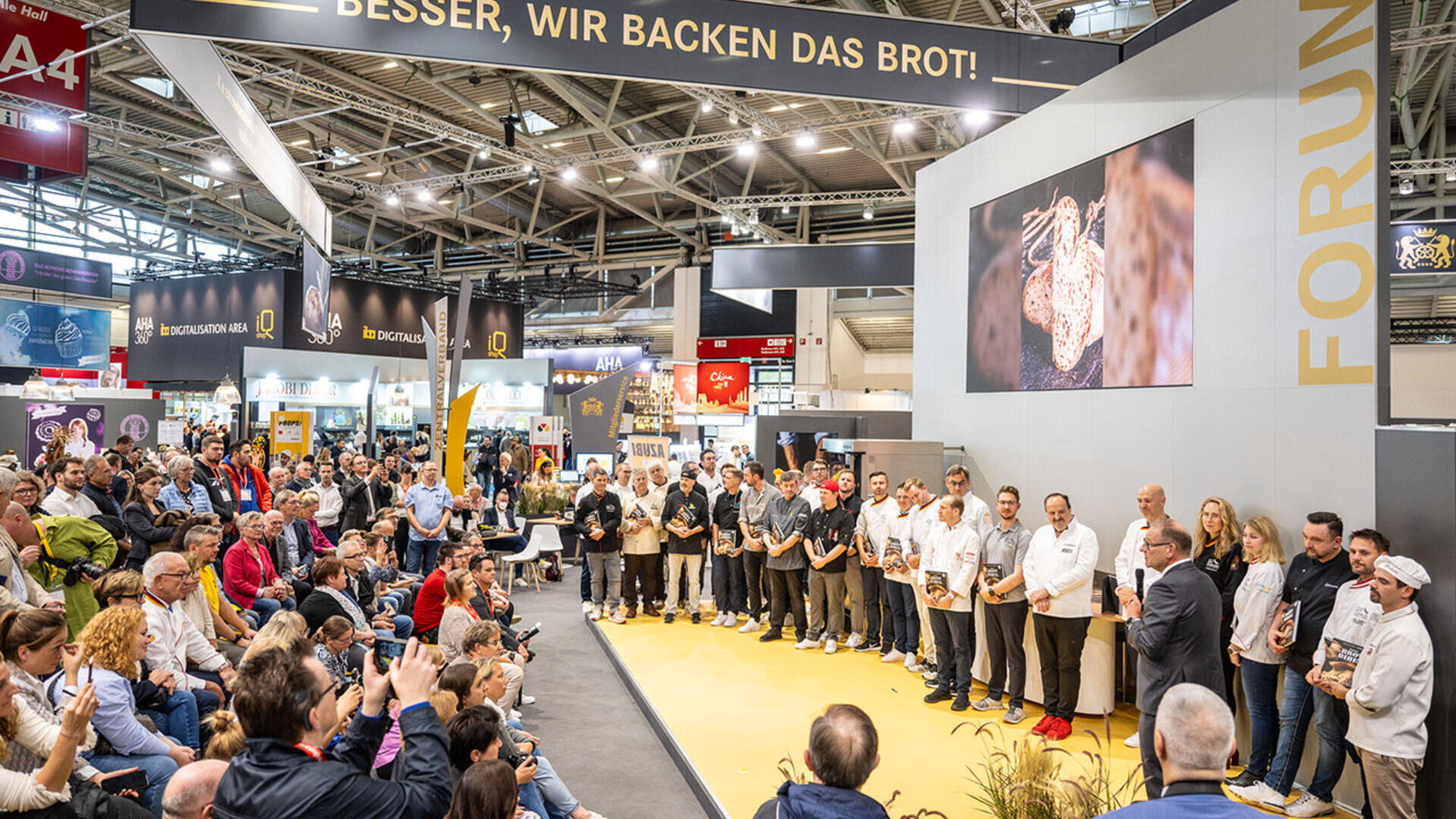A large audience watches a group of people standing on a stage at a trade show or conference, with a screen displaying bread and a sign above reading BESSER, WIR BACKEN DAS BROT! in a spacious exhibition hall.