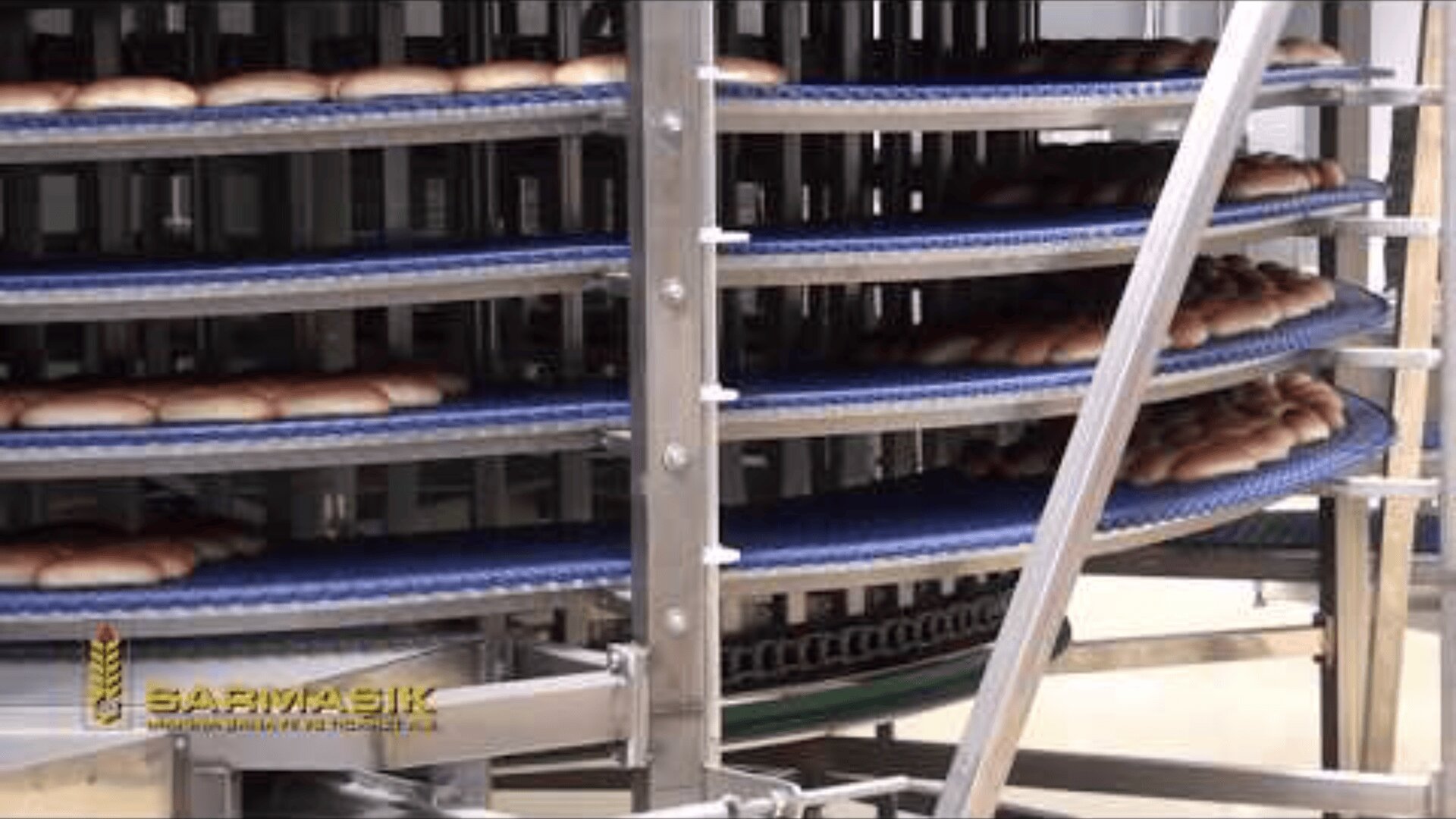 Industrielles Brotkühlband mit mehreren kreisförmigen, vertikal gestapelten blauen Bändern, die Reihen von frisch gebackenem Brot transportieren. In der unteren linken Ecke ist ein Firmenlogo mit Weizen und dem Namen SARMAŞIK zu sehen.