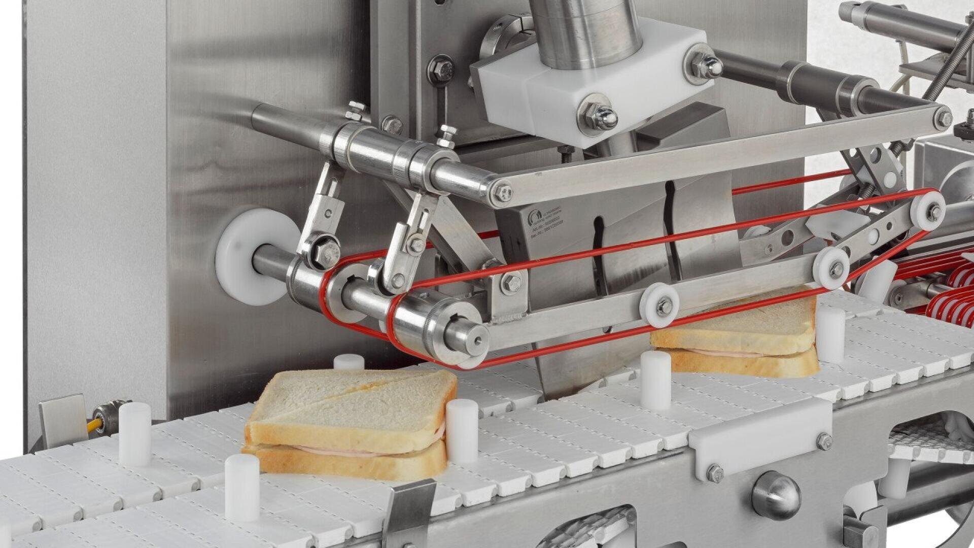 A close-up of a sandwich assembly machine slicing sandwiches in half as they move along a white conveyor belt in a food processing facility. Two sandwiches are visible under the mechanical blade.