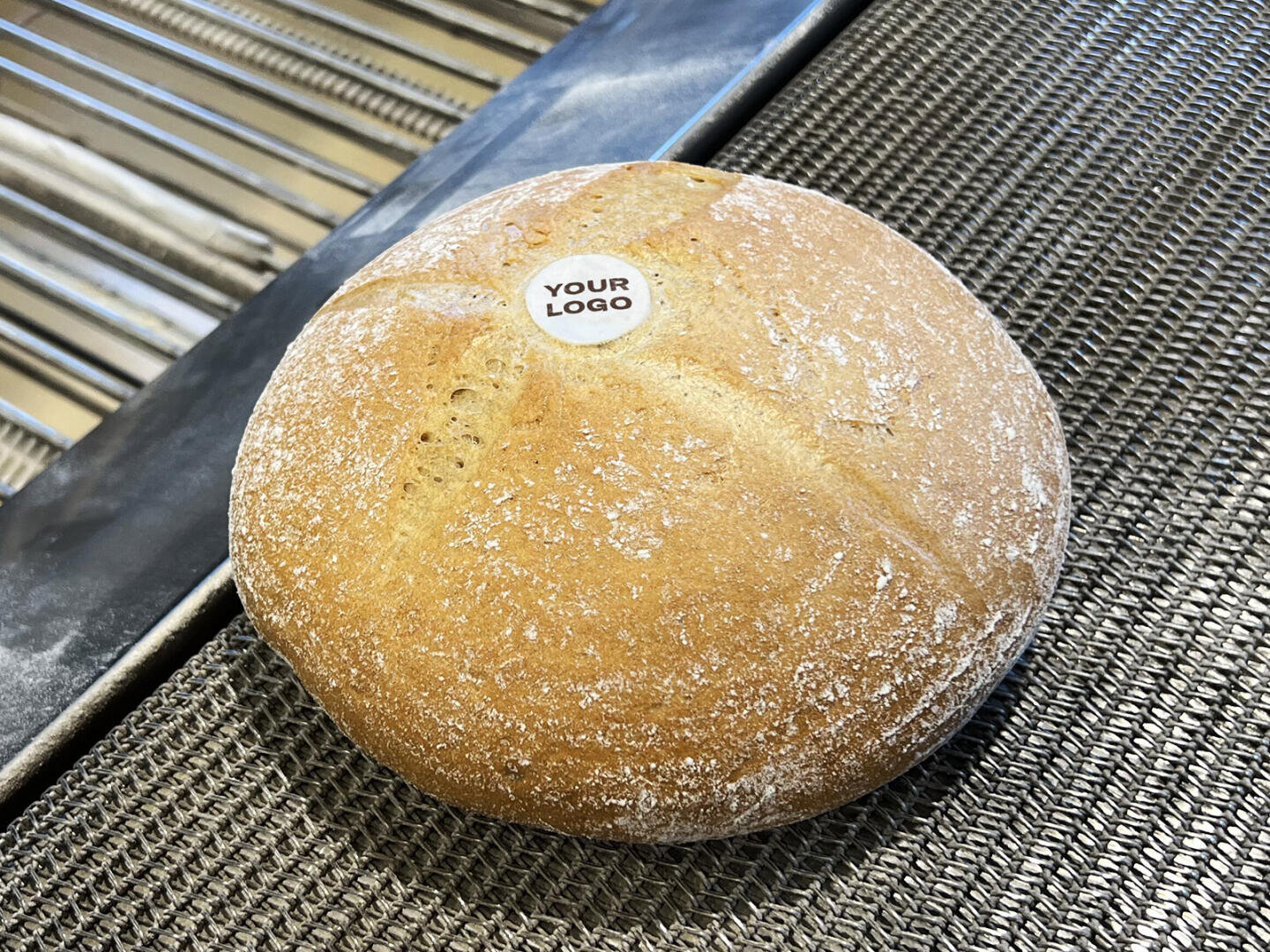 Ein runder Laib Brot mit einem kleinen weißen Aufkleber mit der Aufschrift „IHRES LOGO“ obenauf, der auf einem Metallförderband in einer Bäckerei liegt.