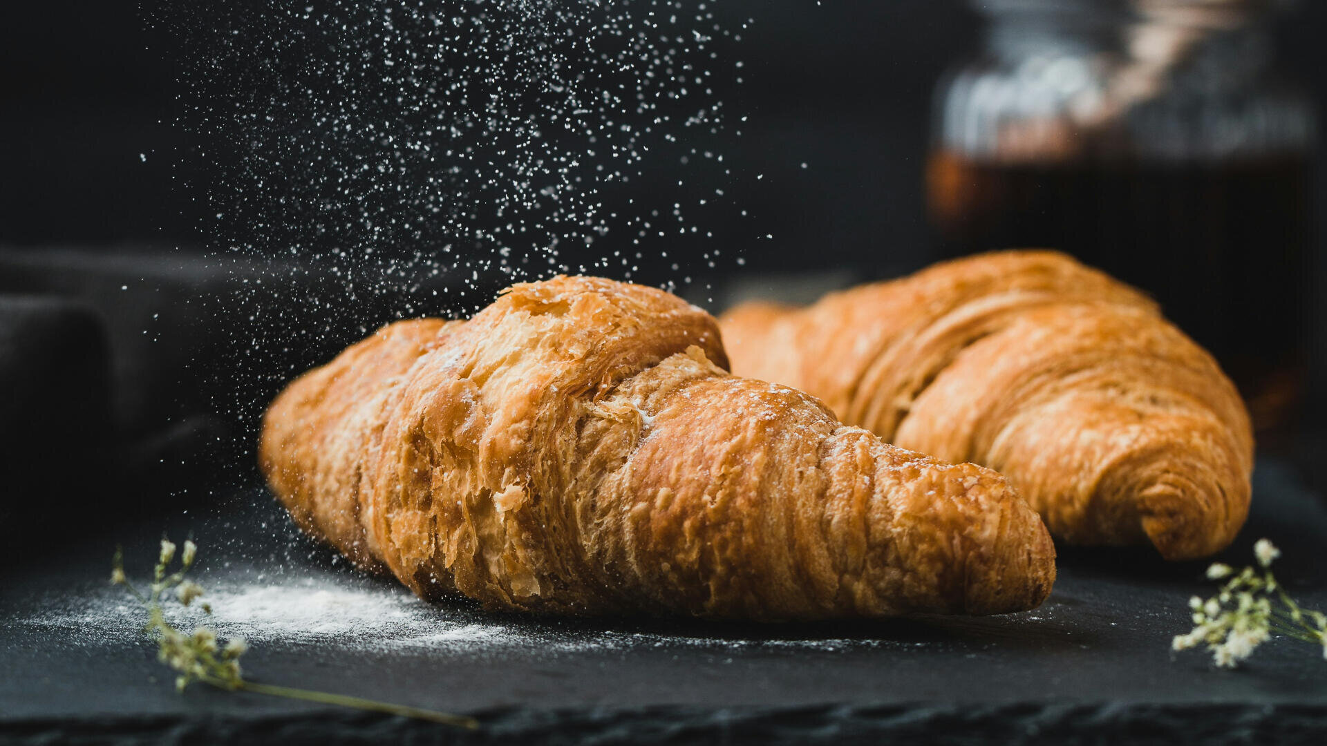 Eine Nahaufnahme von zwei goldenen, flockigen Croissants auf einer dunklen Fläche, über die Puderzucker gestreut wird. Im Hintergrund ist unscharf ein Glas zu sehen, und neben den Croissants stehen ein paar Zweige mit kleinen weißen Blumen.