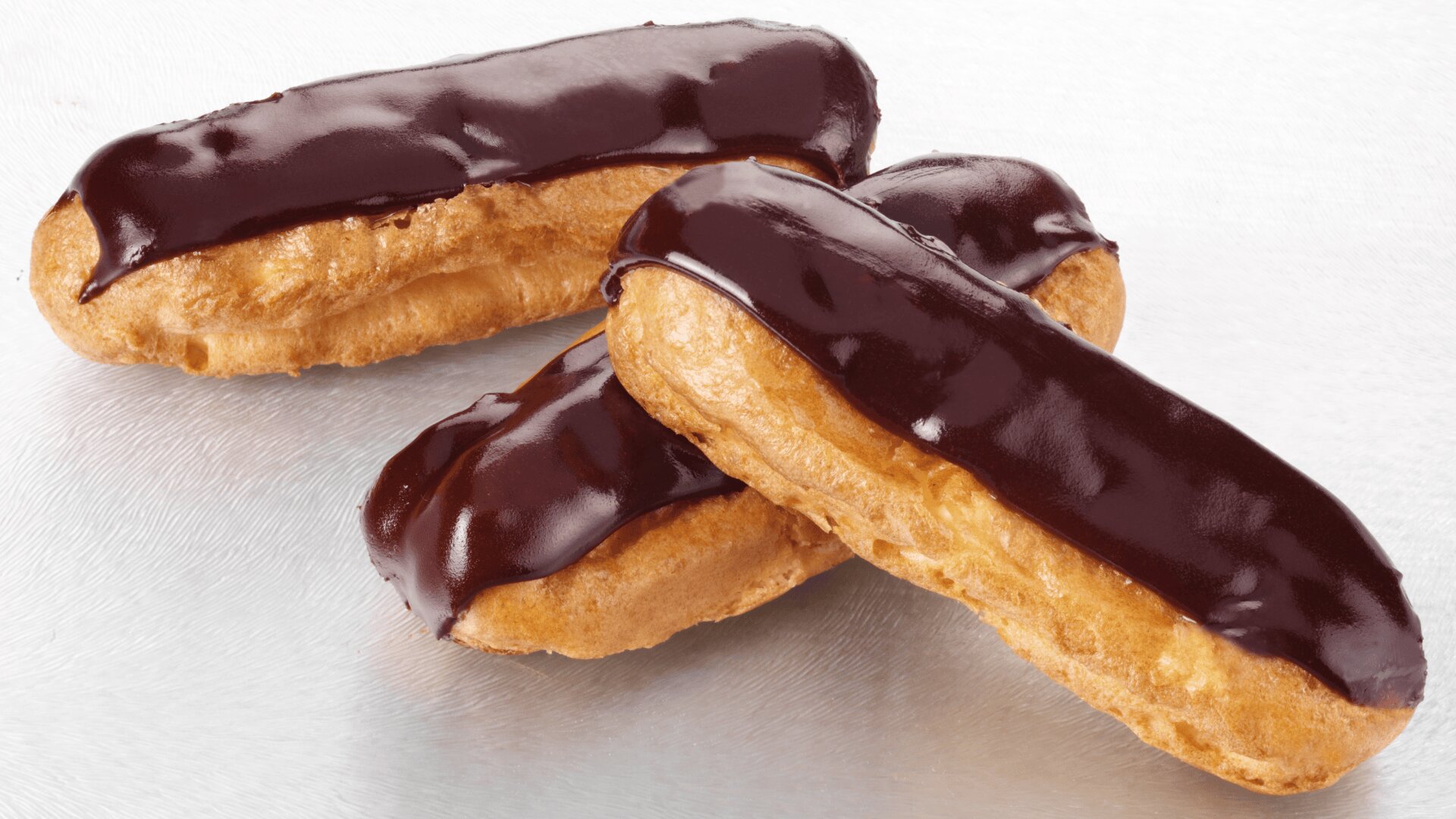Three chocolate eclairs with glossy chocolate icing on top, placed on a light, textured surface. The pastries have a golden-brown choux exterior and appear freshly made.
