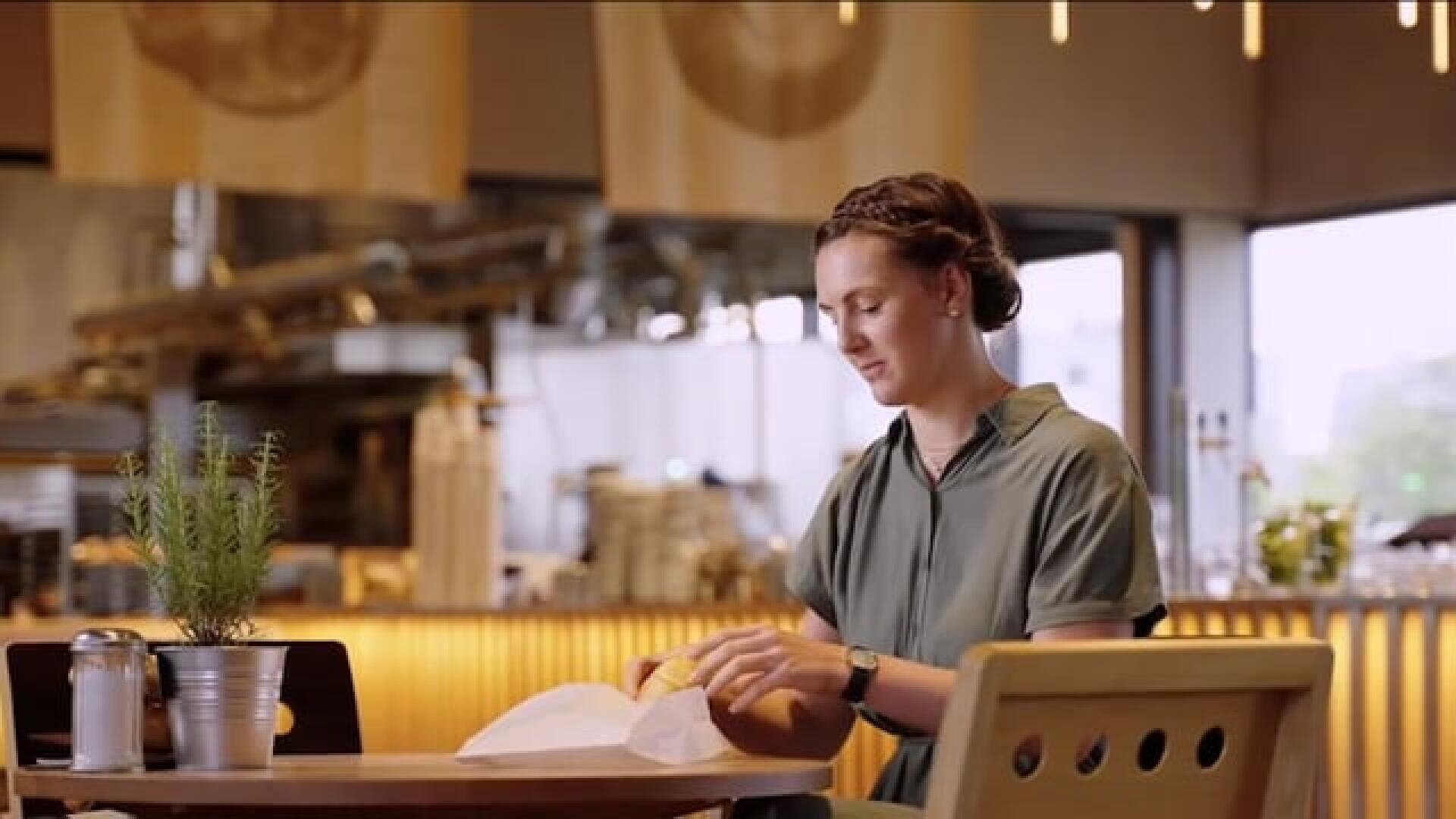 Eine Frau mit geflochtenem Haar sitzt allein an einem Tisch in einem modernen Café und packt ihr Essen aus. Auf dem Tisch befinden sich eine Topfpflanze, ein Salzstreuer und ein Serviettenhalter. Im Hintergrund ist ein heller, offener Küchenbereich zu sehen.