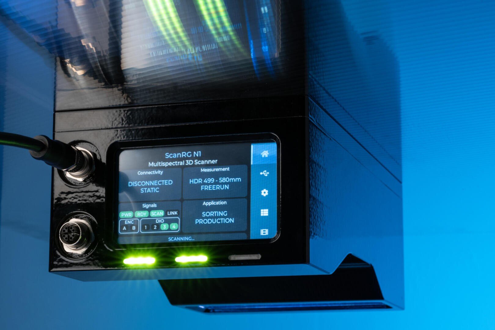Eine Nahaufnahme eines multispektralen 3D-Scanners mit einem Bildschirm, der Statusinformationen und Bedienelemente anzeigt. Das Gerät ist schwarz, verfügt über Anzeigeleuchten und Kabel und steht vor einem blau beleuchteten Hintergrund.