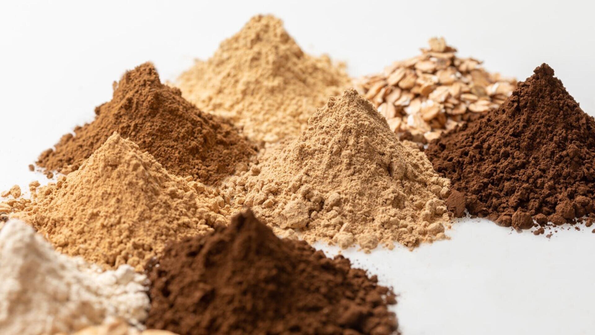 Small piles of various powders in different shades of brown and beige are arranged on a white surface, with a cluster of oat flakes visible in the background.