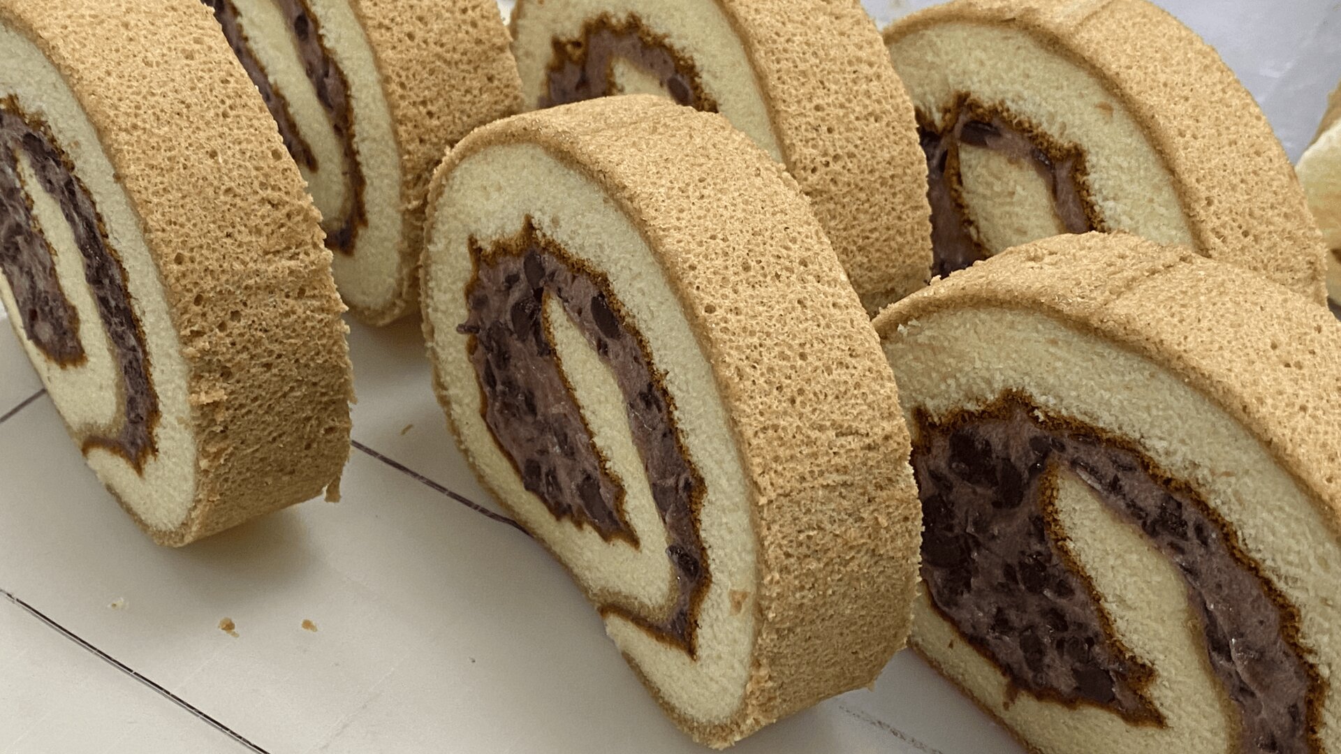 Mehrere Scheiben eines gerollten Biskuitkuchens, der mit einer dunkelroten Bohnenpaste gefüllt ist, sind sauber auf einer hellen Fläche angeordnet. Das spiralförmige Muster von Kuchen und Füllung ist deutlich zu erkennen.