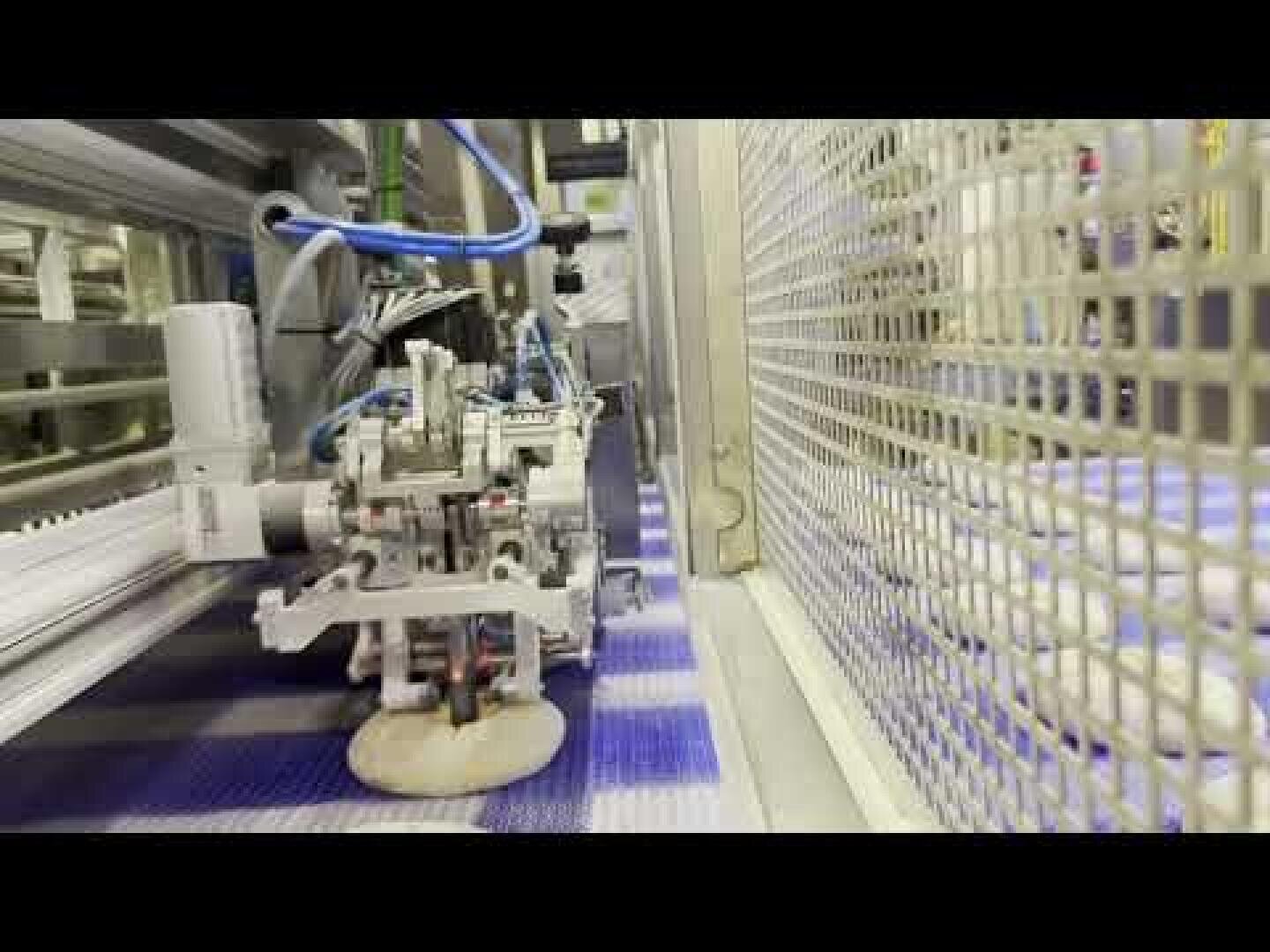In einer Lebensmittelverarbeitungsanlage, die von Metallgeräten und Zäunen umgeben ist, legt eine Robotermaschine ein rundes Stück Teig auf ein Förderband.