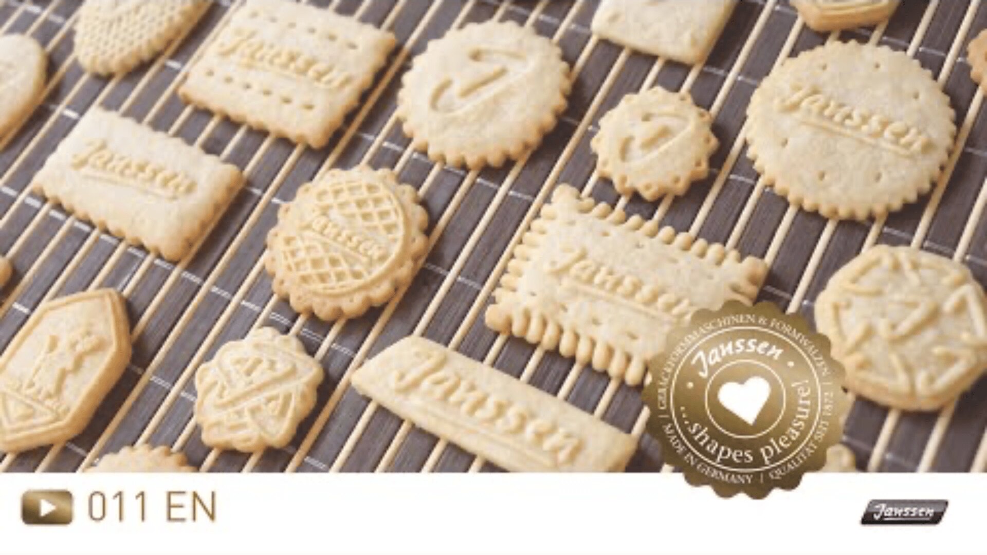 Verschiedene Butterkekse mit unterschiedlichen Formen, Mustern und dem eingeprägten Wort Janssen kühlen auf einem Drahtgestell ab. Auf einem goldenen Siegel in der Ecke steht Janssen Shapes Plaisir!.