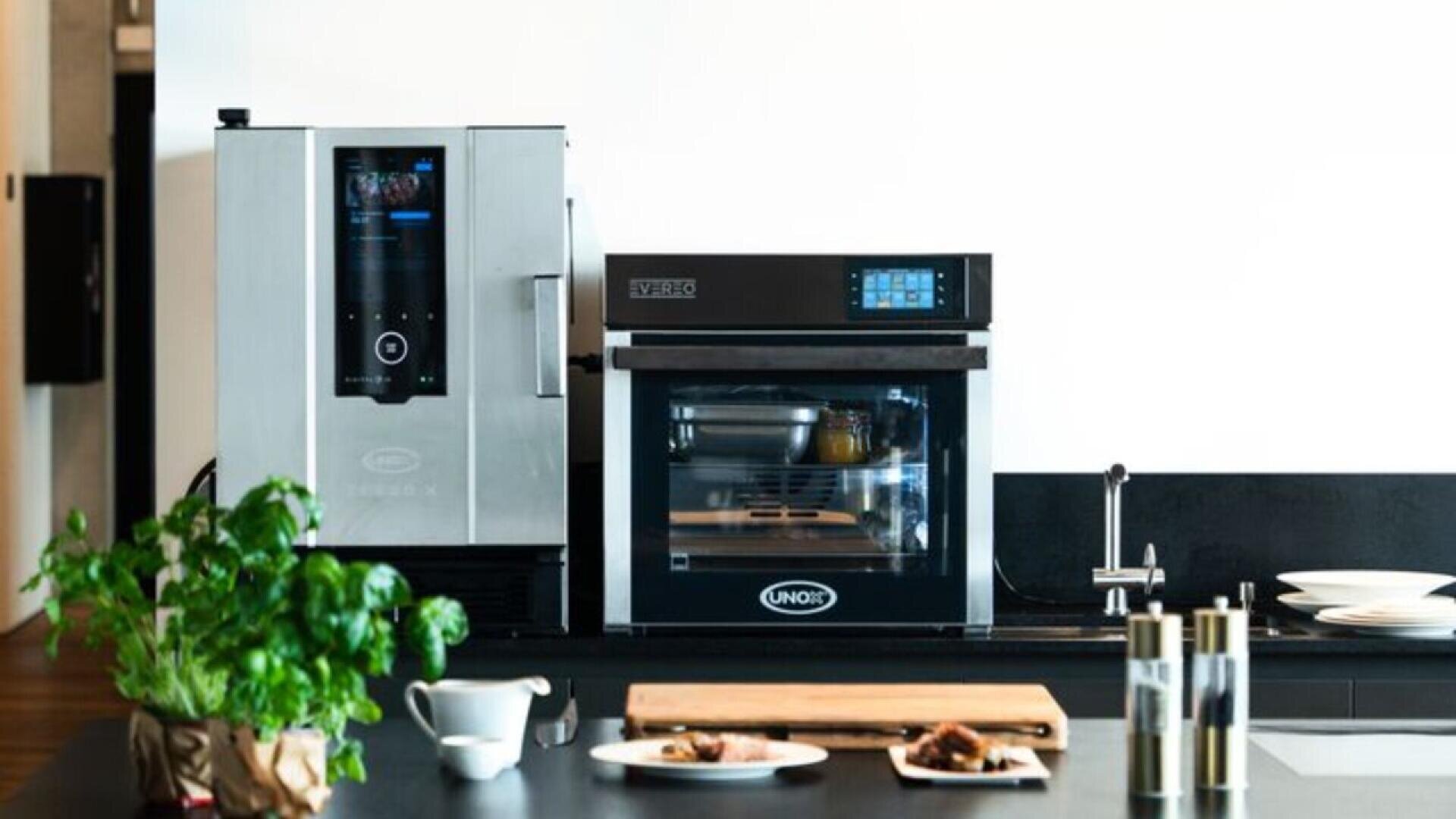 A modern kitchen countertop with a coffee machine, an oven, a wooden cutting board, a mug, herbs in a pot, salt and pepper shakers, and plates with food against a white wall.
