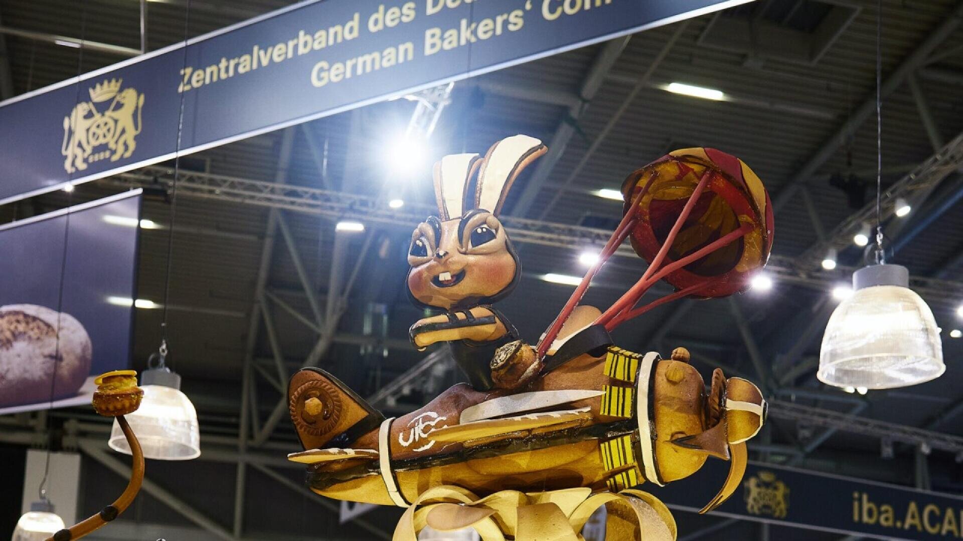 A large, intricate bread sculpture of a rabbit wearing a pharaoh headdress is displayed at an event, with a sign above reading “German Bakers’ Com”. The sculpture is detailed and surrounded by bread and baking-themed elements.