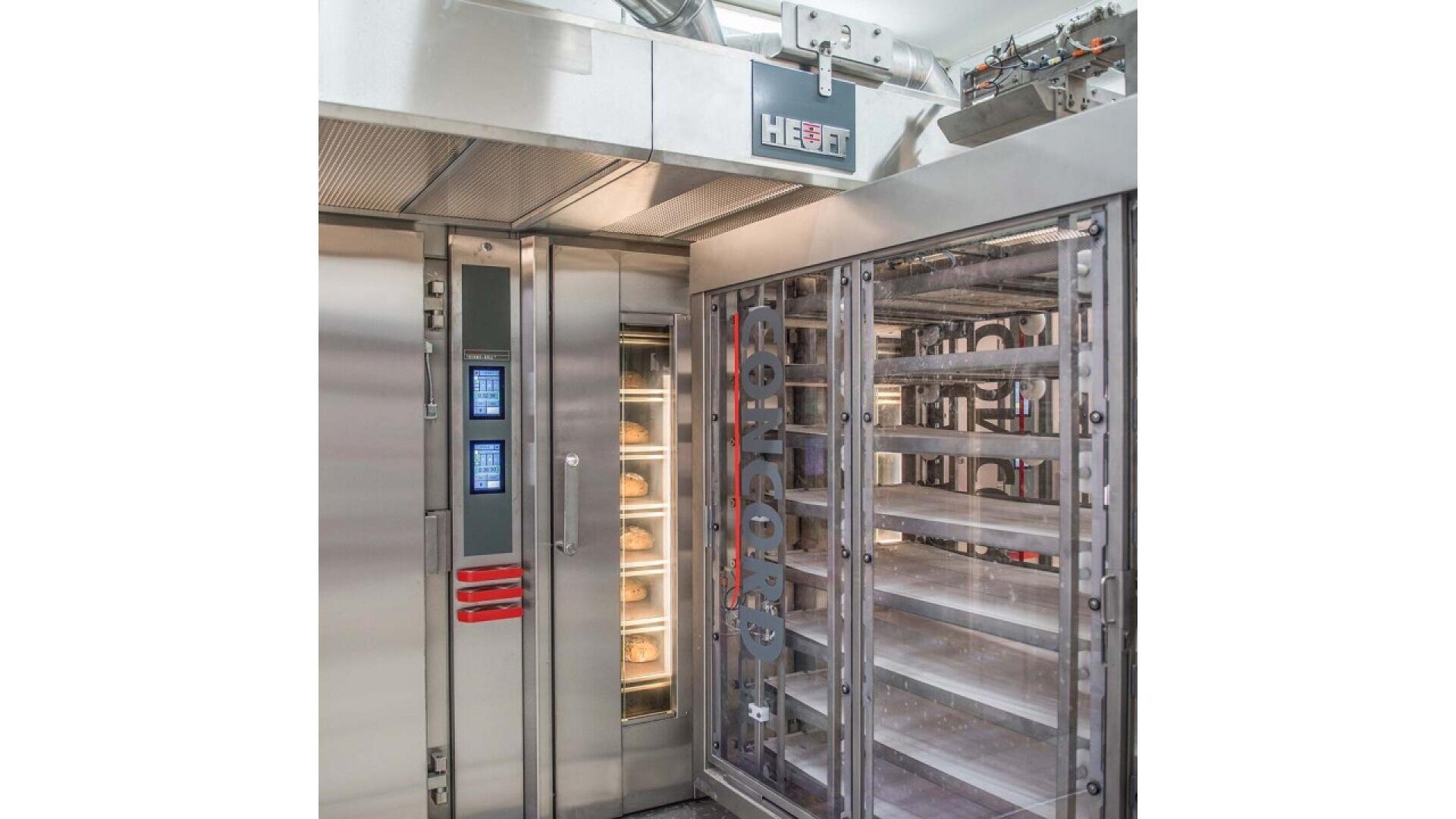 A commercial bakery oven with digital controls and trays of bread inside, next to an empty rack ready for loading, in a clean industrial kitchen setting.