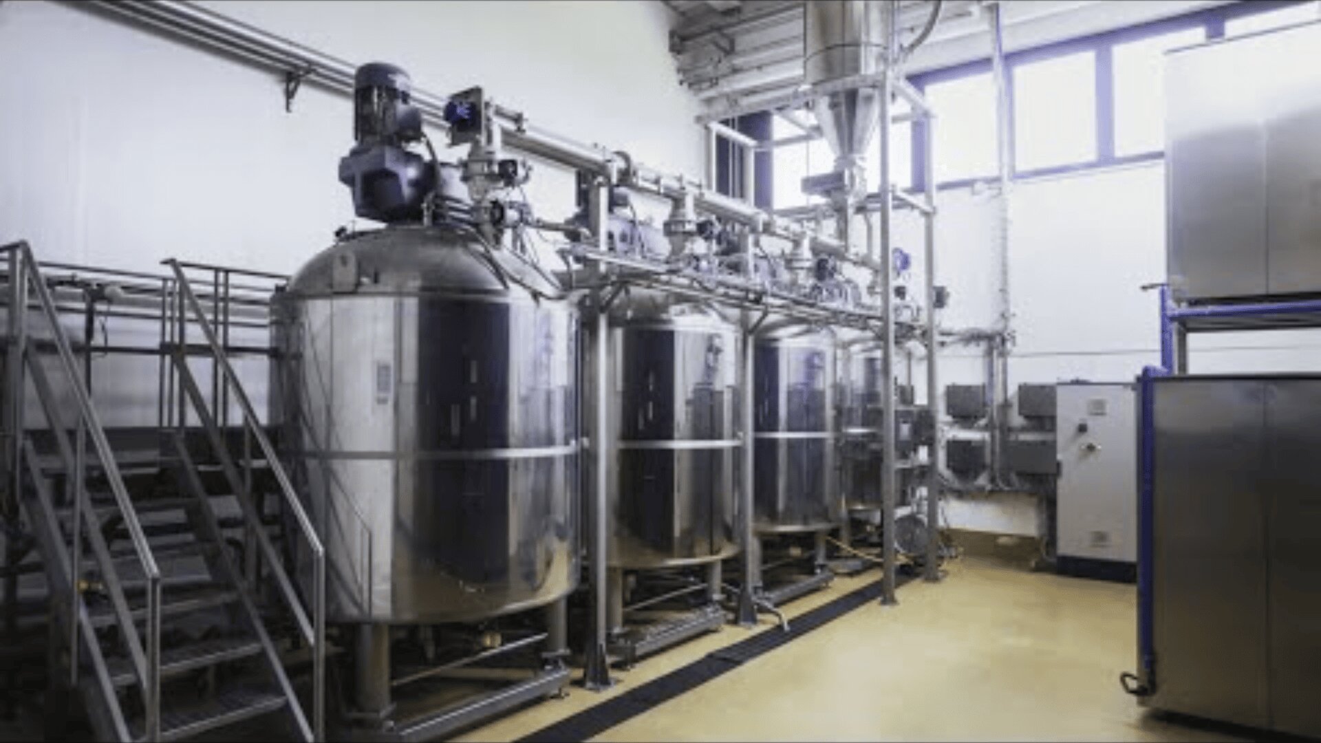 Eine moderne Industrieanlage mit drei großen Edelstahltanks, Metalltreppen und Rohren entlang der Wände, die zur Herstellung oder Verarbeitung genutzt wird und durch das natürliche Licht der hohen Fenster hell erleuchtet wird.