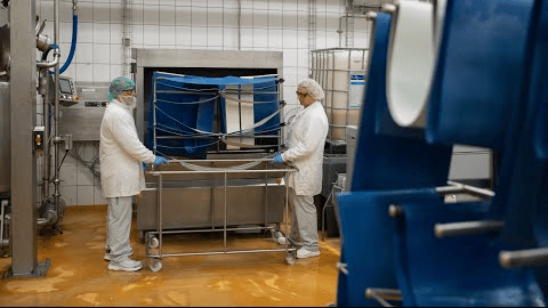 Two workers in white coats, hairnets, and gloves stand in a clean, tiled industrial facility, handling equipment with blue components and large metal containers. The floor is orange-brown.