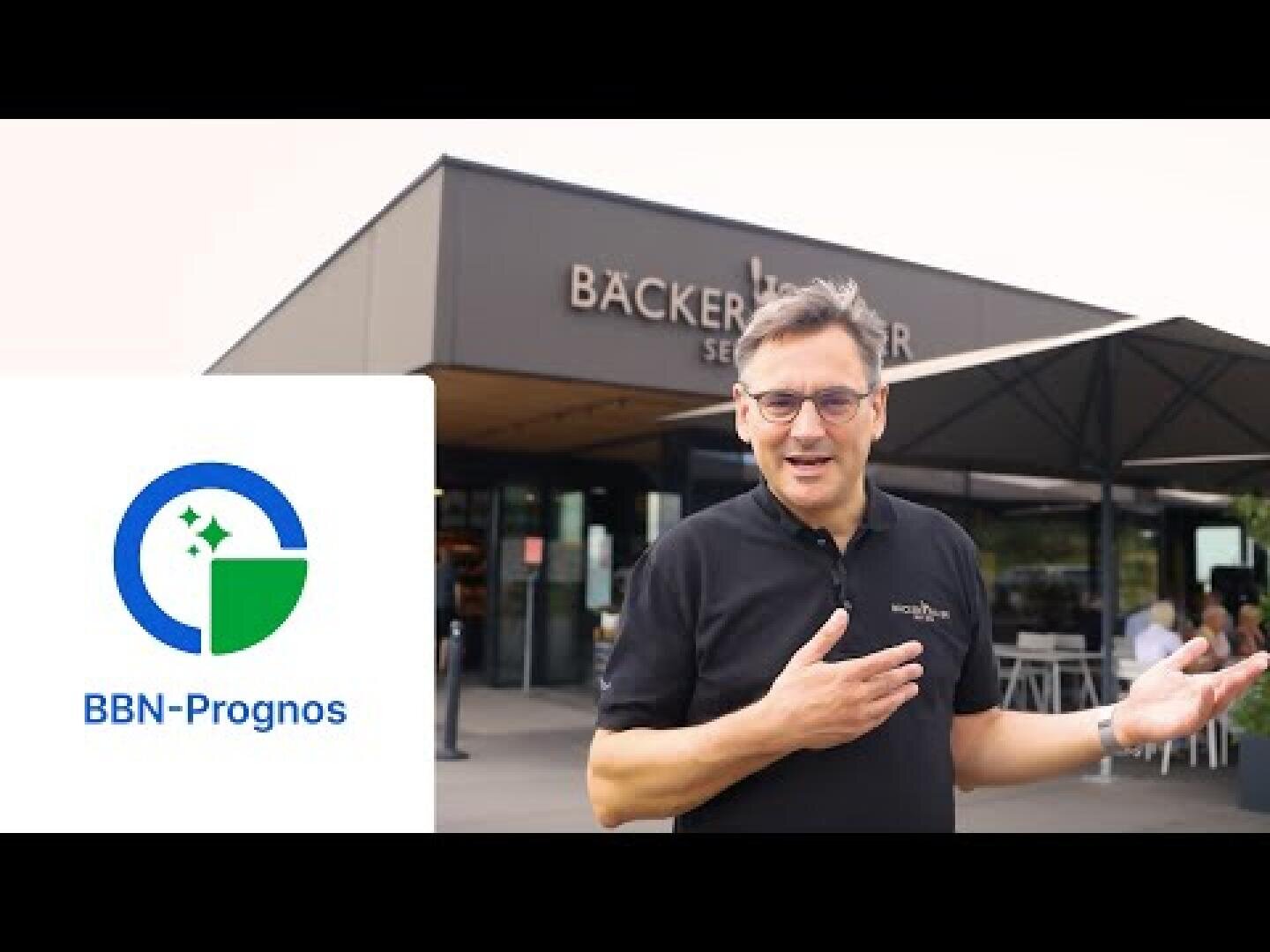 Ein Mann im schwarzen Hemd steht gestikulierend vor einem Bäckerei-Café. Über dem Eingang steht „BÄCKER“. Links sind das Logo und der Text von BBN-Prognos auf weißem Hintergrund zu sehen.