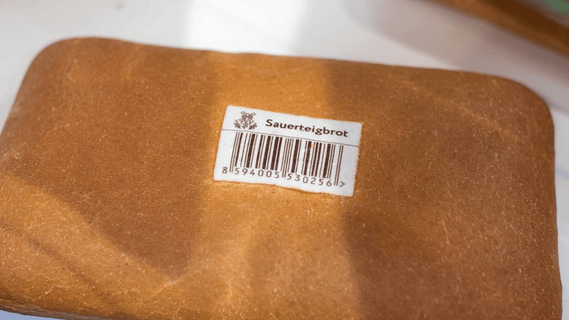 A rectangular loaf of bread with a label reading Sauerteigbrot and a barcode is placed on a white surface.
