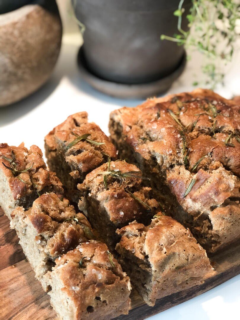Close-up of sliced homemade focaccia bread on a wooden board, topped with rosemary and sea salt, with blurred potted plants in the background.